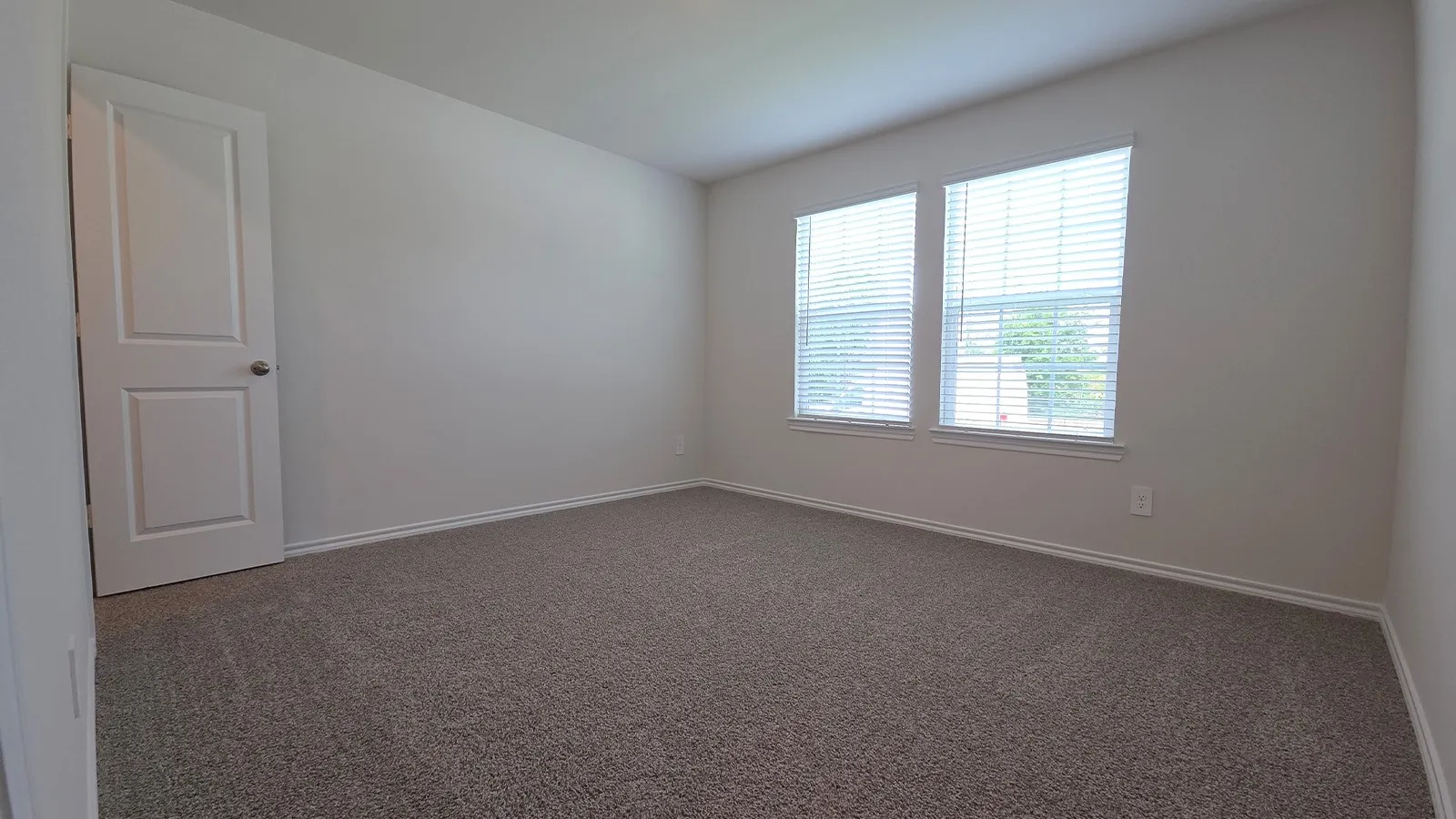 Spacious room featuring neutral wall tones, gray carpeting, dual windows with blinds, and white trim