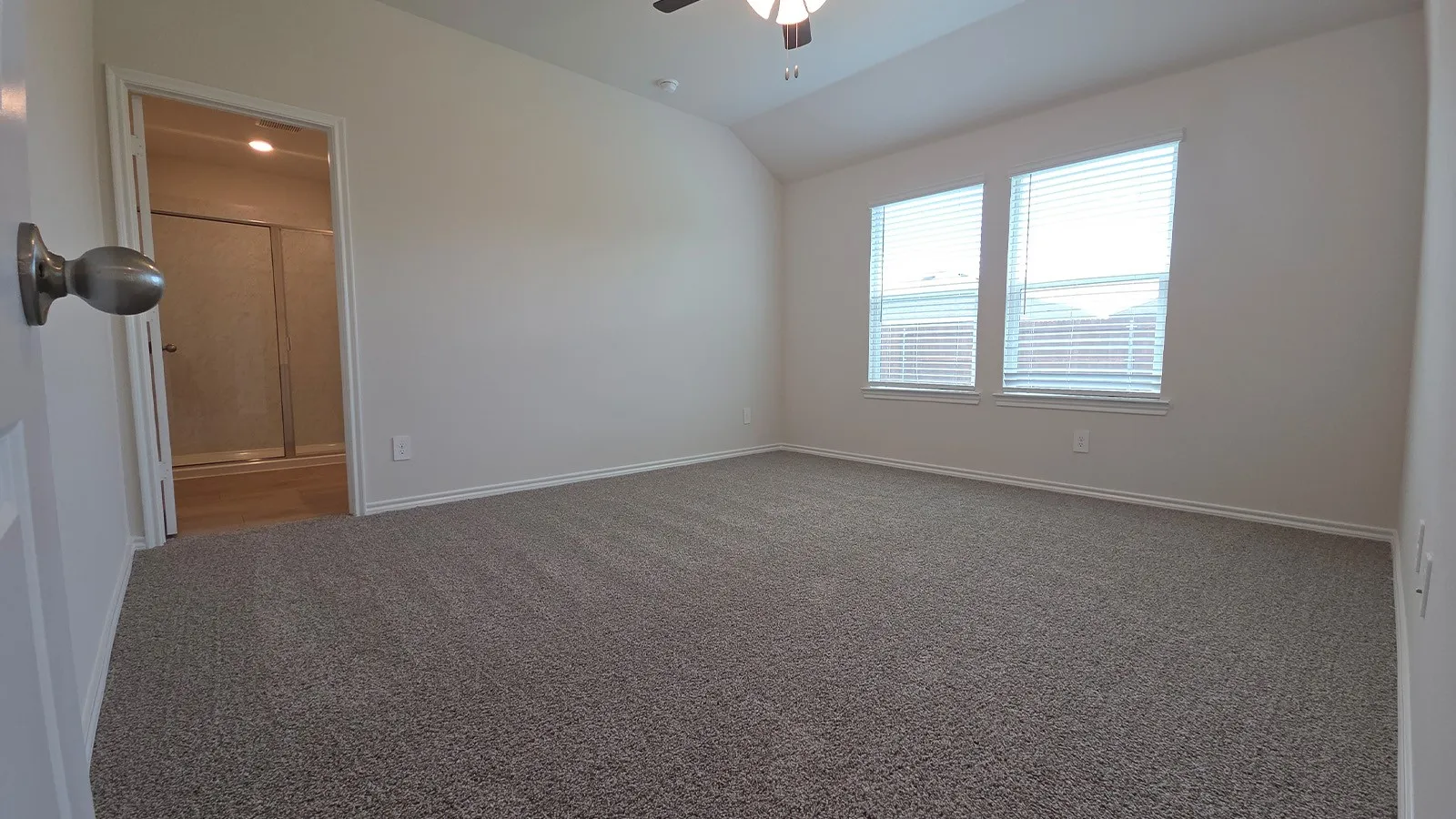 Carpeted bedroom featuring two windows with blinds, a ceiling fan, and a doorway opening to a bathroom with a glass-enclosed shower