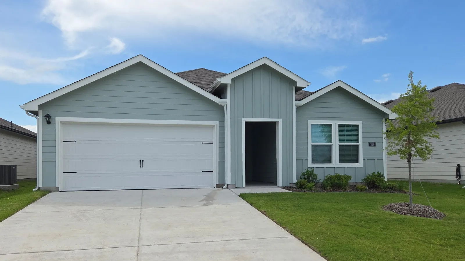 Exterior featuring light blue horizontal siding, vertical board and batten accents, a two-car garage with decorative hardware, a covered entry, and a manicured lawn with a young tree