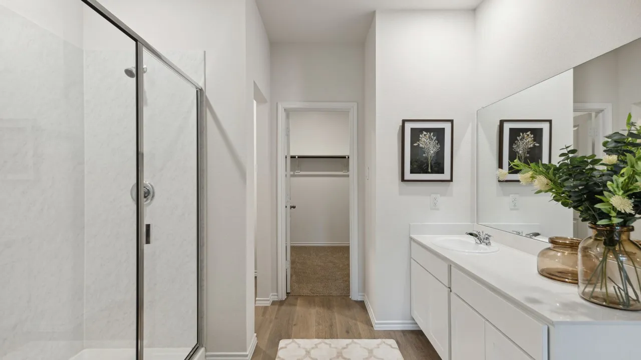 Bathroom featuring a glass-enclosed shower, dual vanity with undermount sinks, expansive mirror, wood-finish flooring, and a walk-in closet beyond