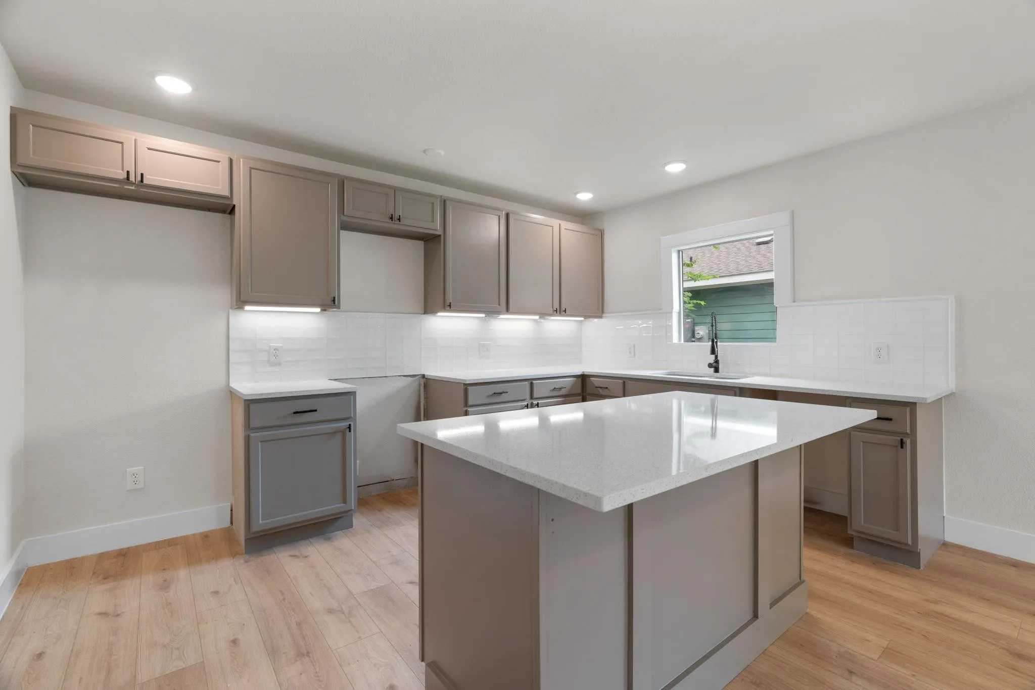 Contemporary kitchen featuring an island with a light-toned countertop, shaker-style cabinetry, subway tile backsplash, under-cabinet lighting, and wood-finish flooring