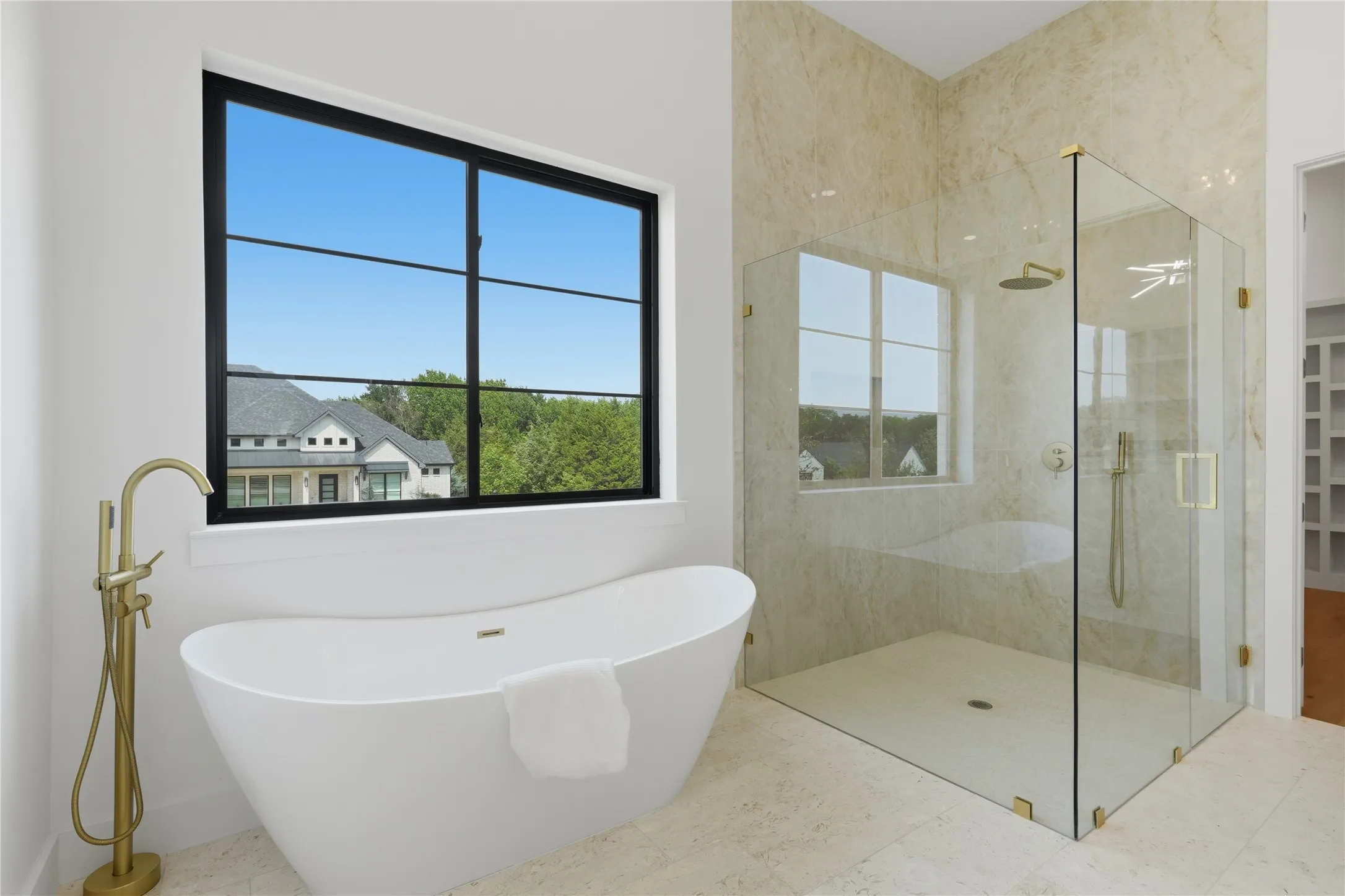 Freestanding soaking tub with a floor-mounted brass-finish faucet, accompanied by a spacious glass-enclosed shower featuring a brass-finish rainfall showerhead and handheld sprayer