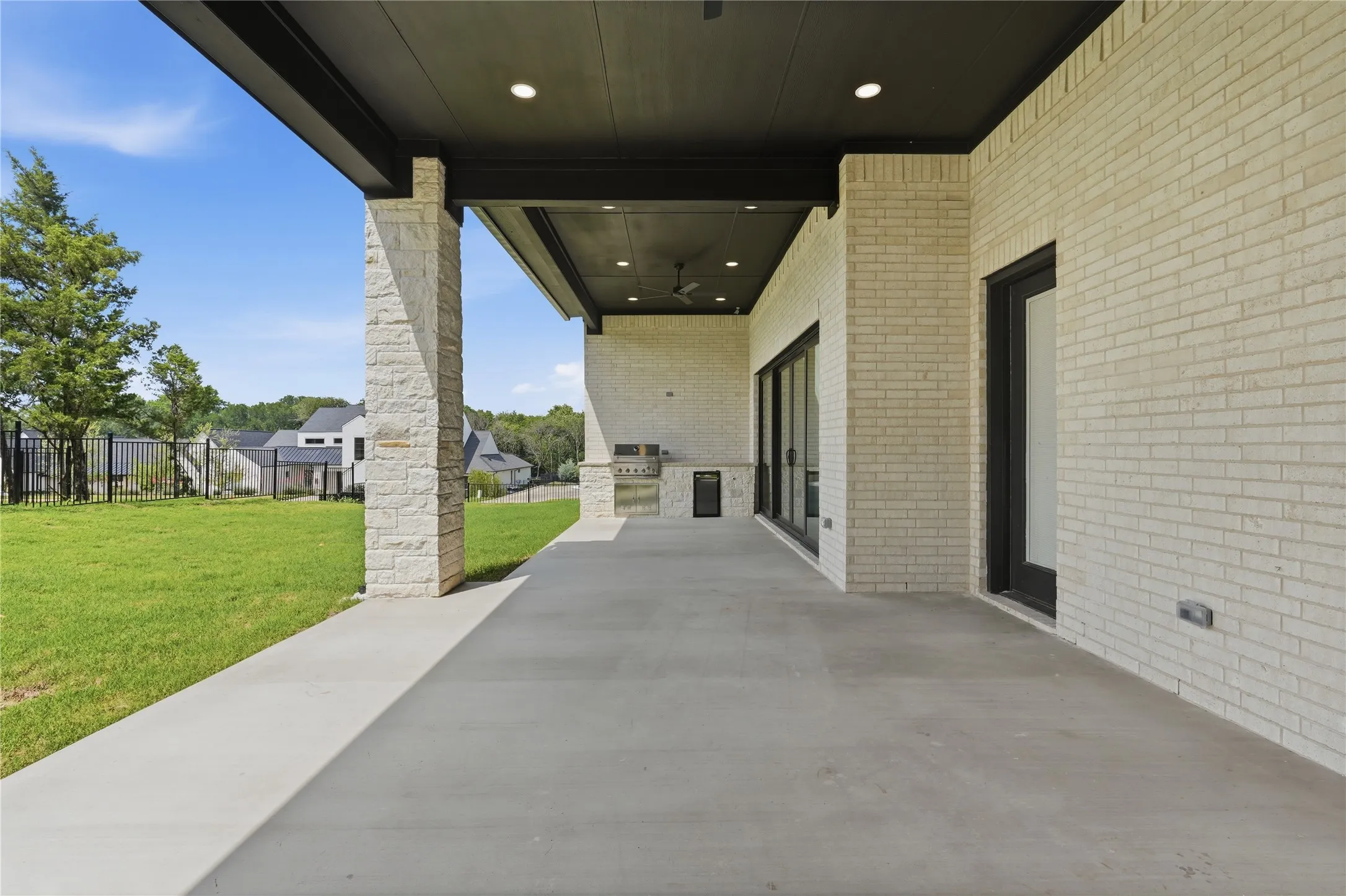 Covered outdoor patio featuring light-toned brick exterior, stone-clad pillars, concrete flooring, and a built-in outdoor kitchen with a grill