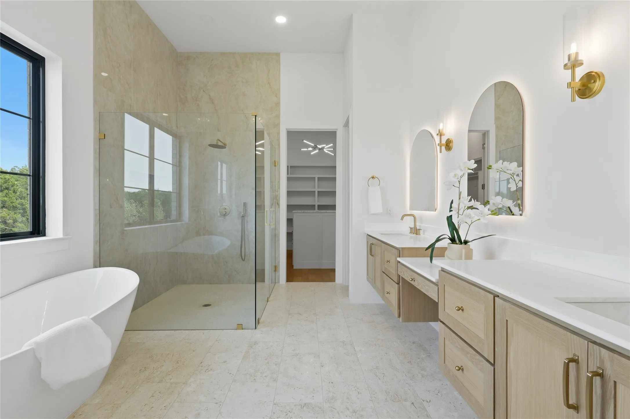 Contemporary bathroom featuring a freestanding soaking tub, a spacious glass-enclosed shower with tile surround, dual floating vanities with light wood-finish cabinetry, and arched illuminated mirrors