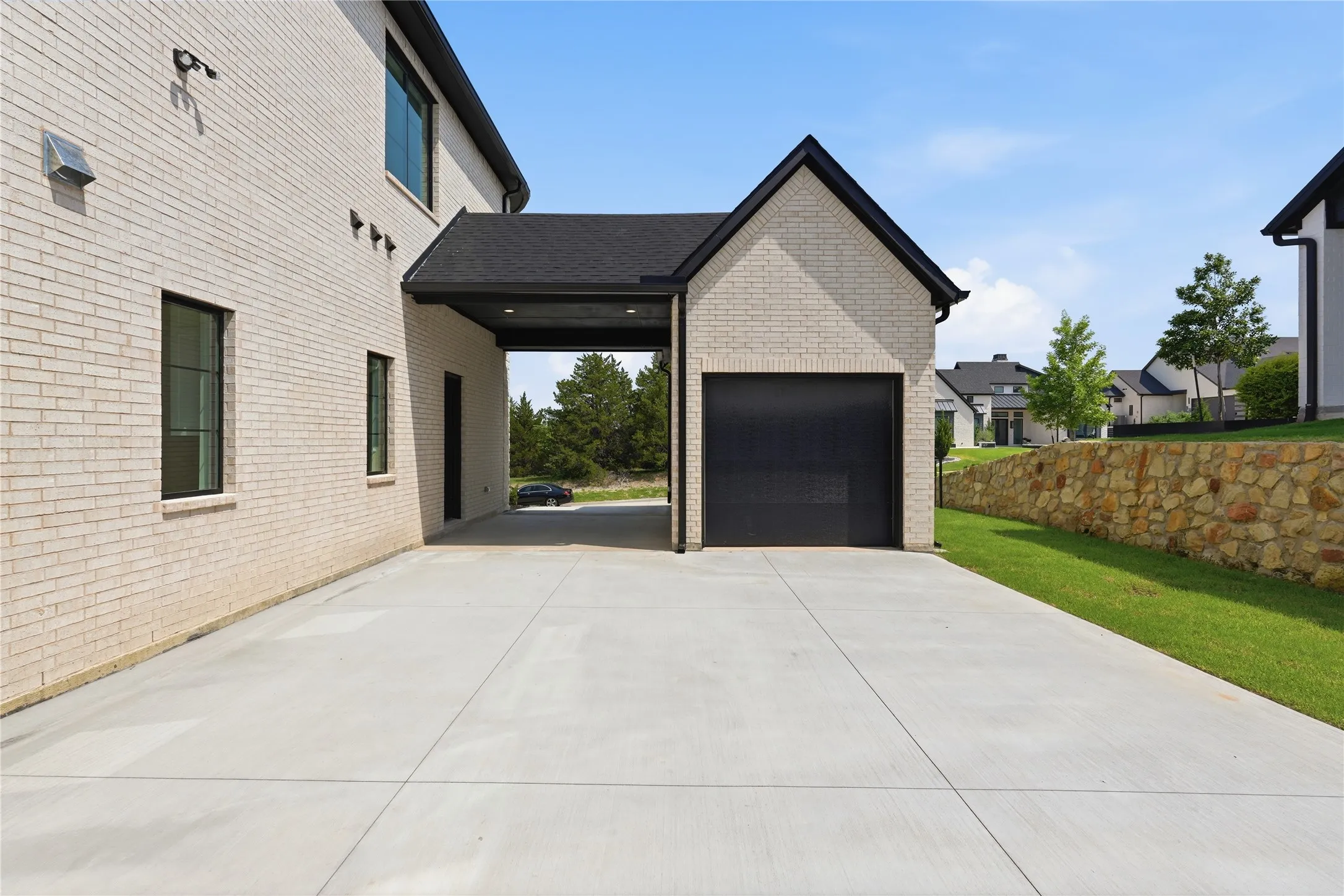 Attached two-car garage with a matte black door, concrete driveway, light brick exterior, dark window frames, and a stone retaining wall