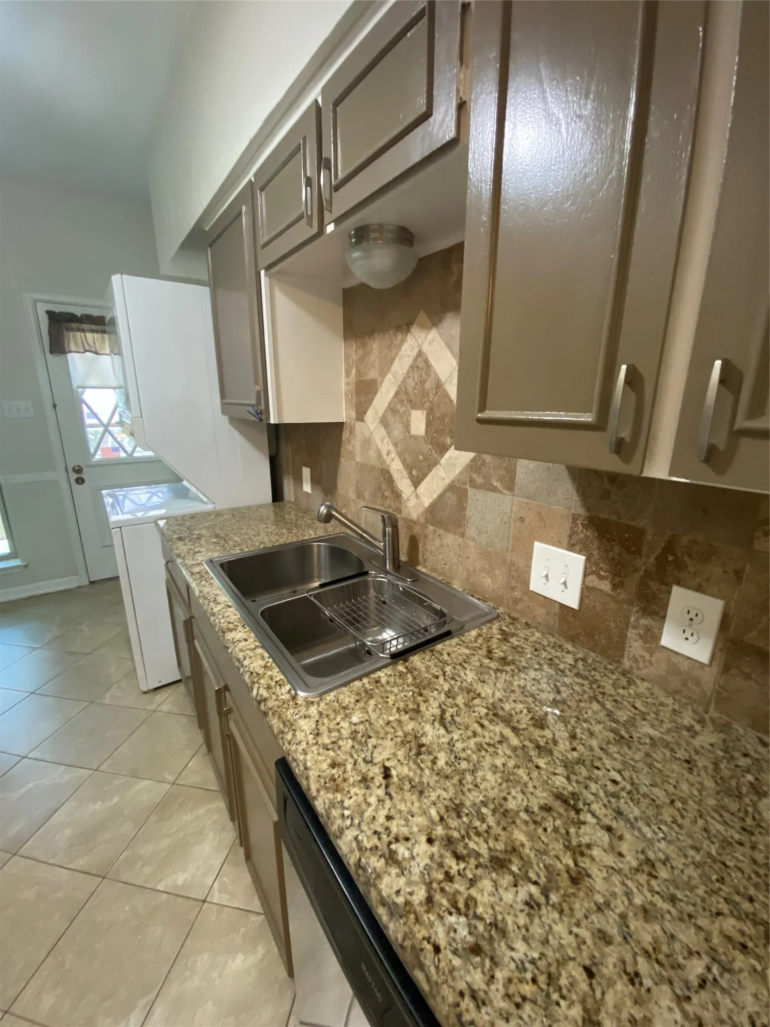 Granite-finish countertops, stainless steel double basin sink, and tile backsplash with decorative inset