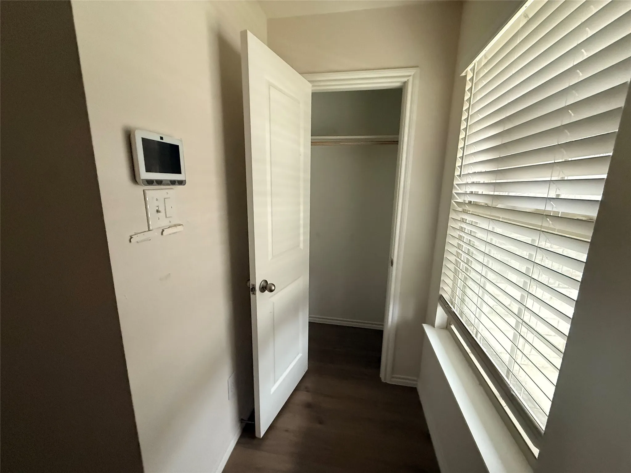 Interior space featuring wood-finish flooring, a white paneled door with a brushed nickel knob, white window blinds, and a built-in wall panel device
