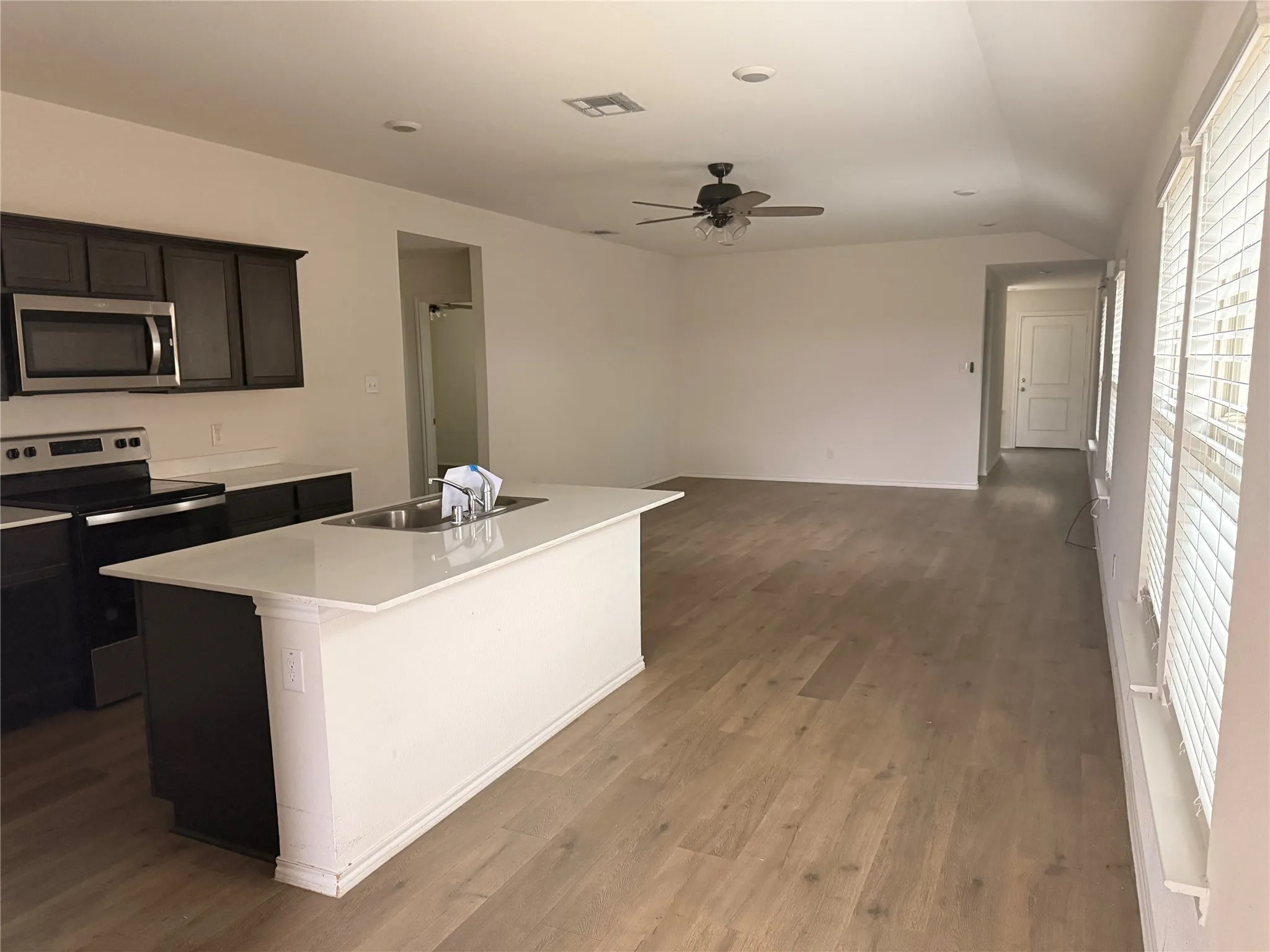 Open-concept living space featuring wood-finish flooring, a kitchen island with integrated sink, dark wood cabinetry with stainless steel appliances, and multiple windows with blinds