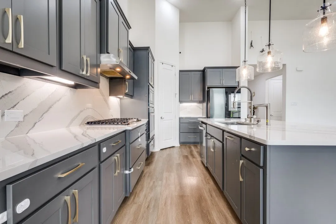 Open-concept kitchen featuring gray shaker cabinetry, gold-toned hardware, white countertops with integrated backsplash, wood-finish flooring, and a central island with a stainless steel sink and modern pendant lighting