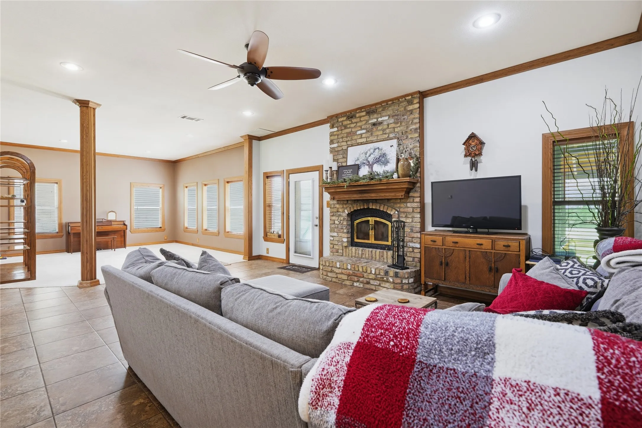 Spacious living area featuring a brick-surround fireplace with a wood mantel, tile flooring, recessed lighting, a ceiling fan, and multiple windows with blinds