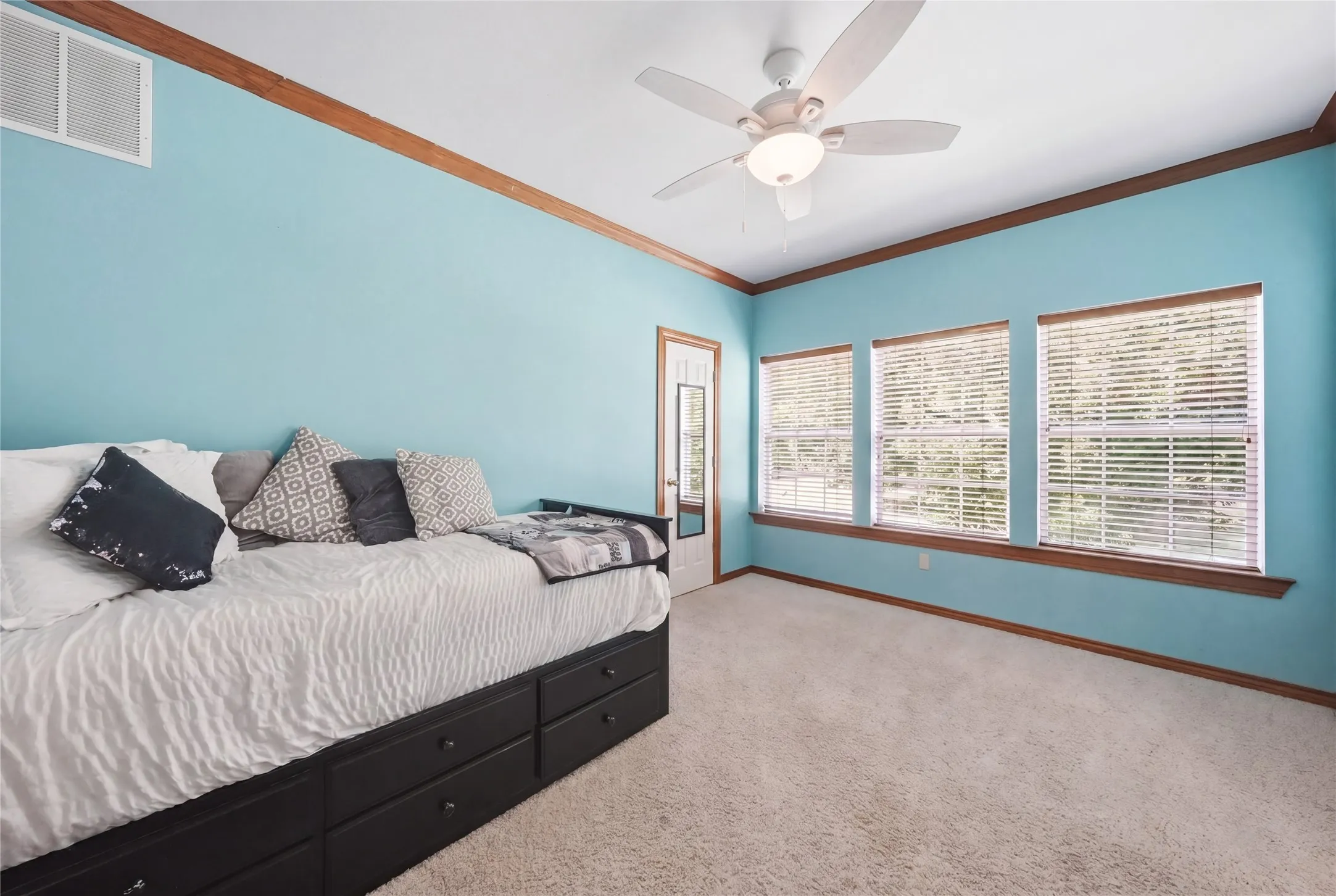 Room featuring light blue walls with wood trim, a built-in ceiling fan with light, three windows with blinds, and light-colored carpeting