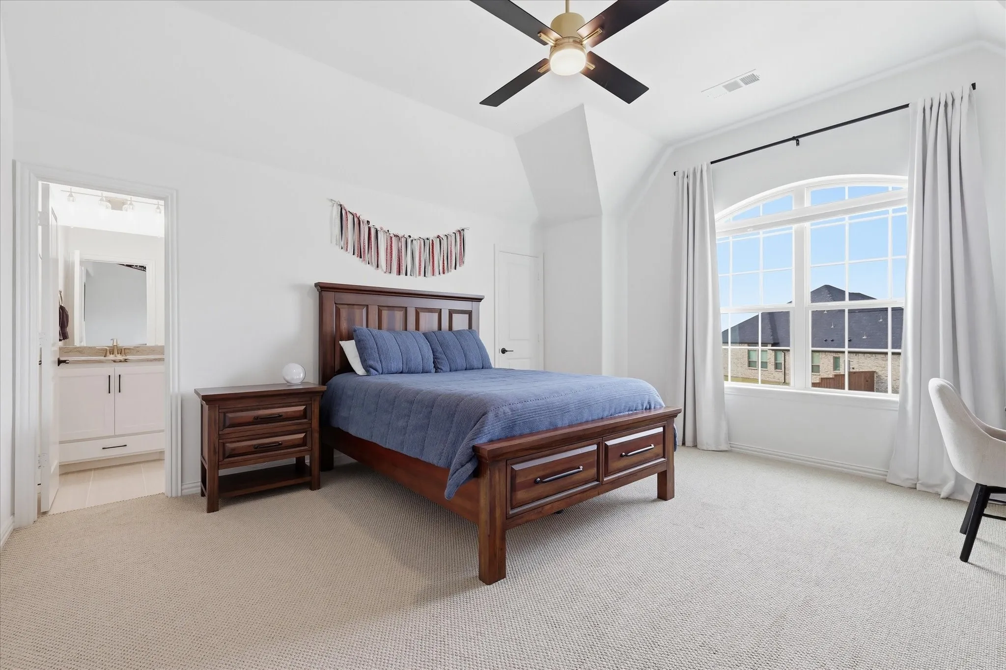 Carpeted bedroom featuring a large arched window, a ceiling fan with integrated lighting, and a white vanity with a gold-toned faucet in the en suite bathroom