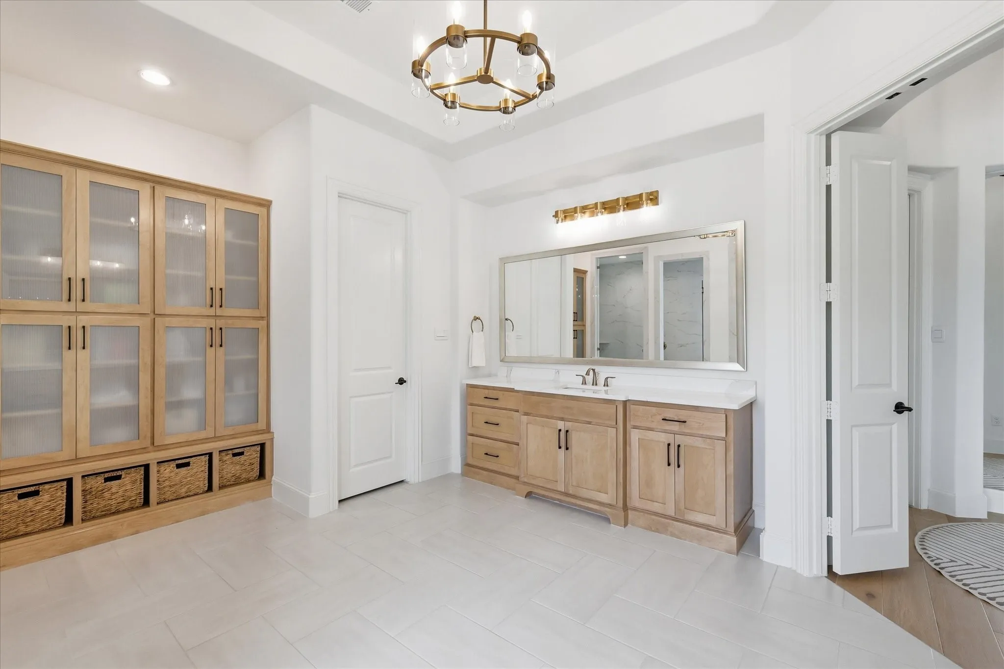 Spacious interior featuring a large wood-finish vanity with a light-colored countertop and double sinks, a substantial built-in storage unit with glass-front cabinetry and base shelving, light-toned tile flooring, and a contemporary chandelier