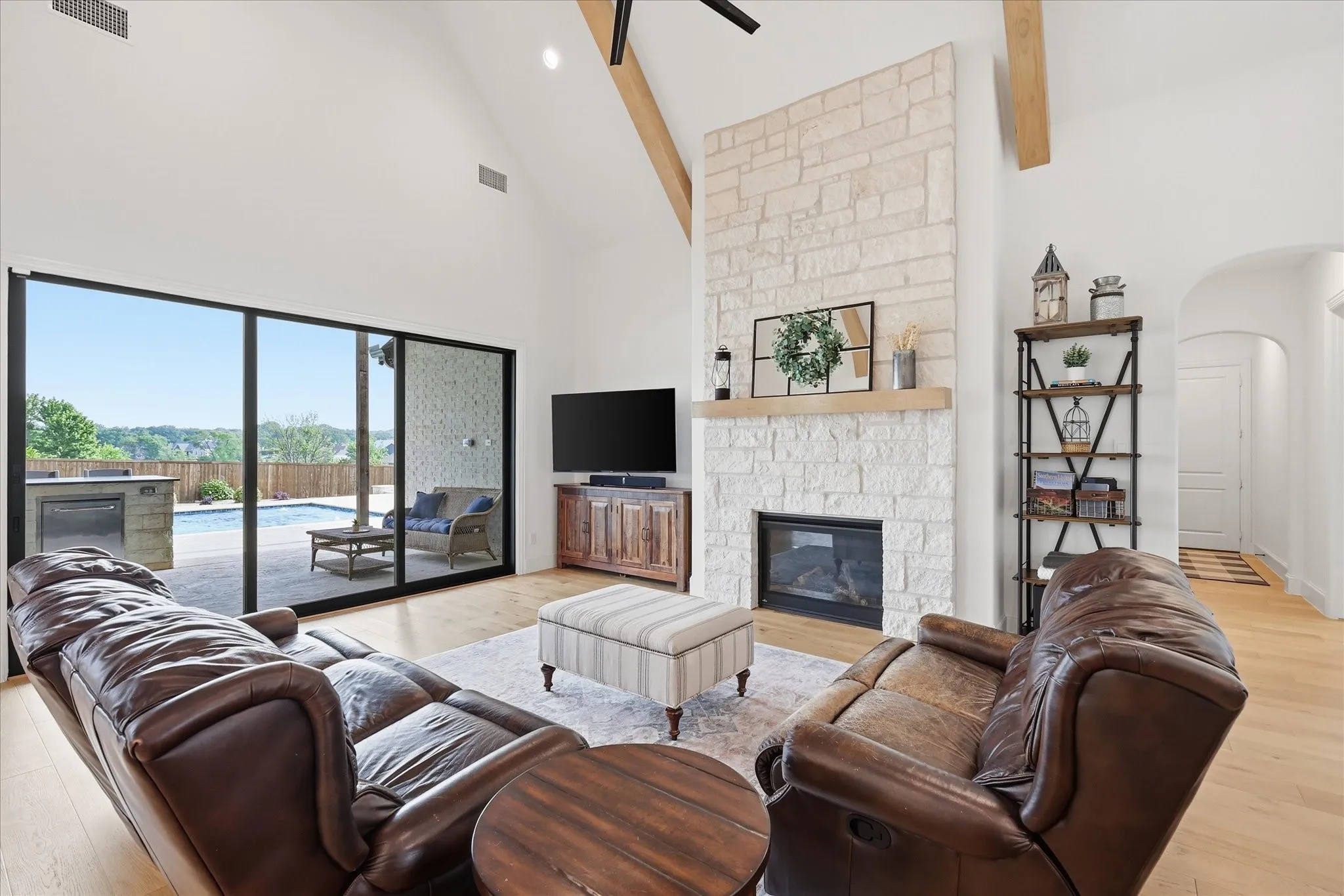 Vaulted ceiling with exposed wood beams, stone fireplace, wood-finish flooring, and expansive sliding glass doors leading to the outdoor space with a built-in grill and swimming pool