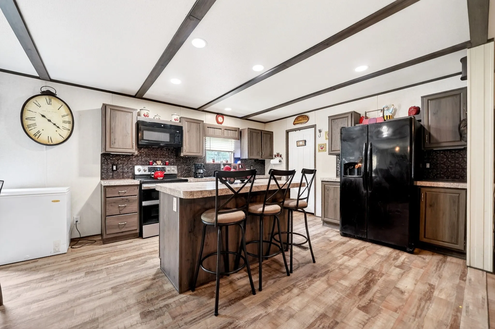 Spacious kitchen featuring an island with breakfast bar seating, dark wood-finish cabinetry, black appliance suite, textured backsplash, and wood-finish flooring