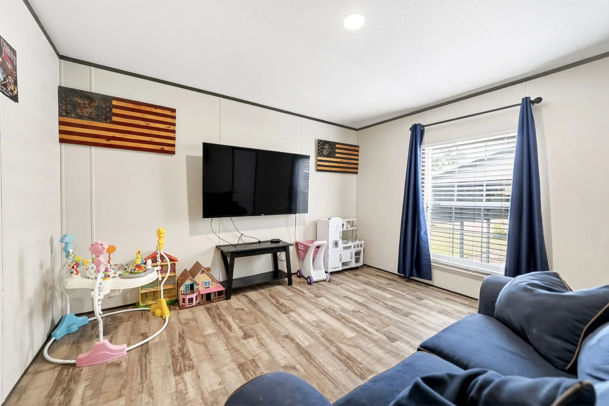 Spacious living area featuring wood-finish flooring, white paneled walls, and a large window with blinds