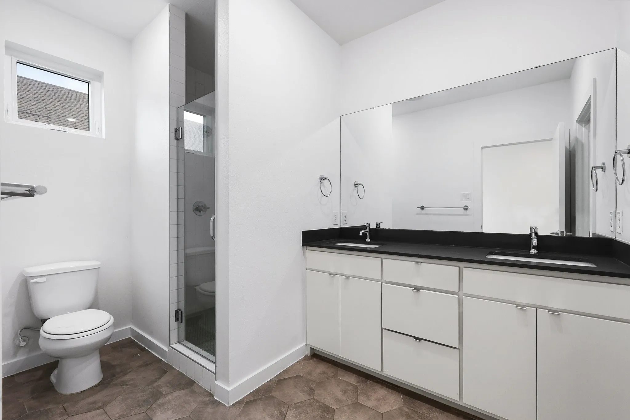 Bathroom featuring a dual vanity with a dark countertop and integrated sinks, a large frameless mirror, a glass-enclosed shower, a white toilet, and hexagonal floor tiling