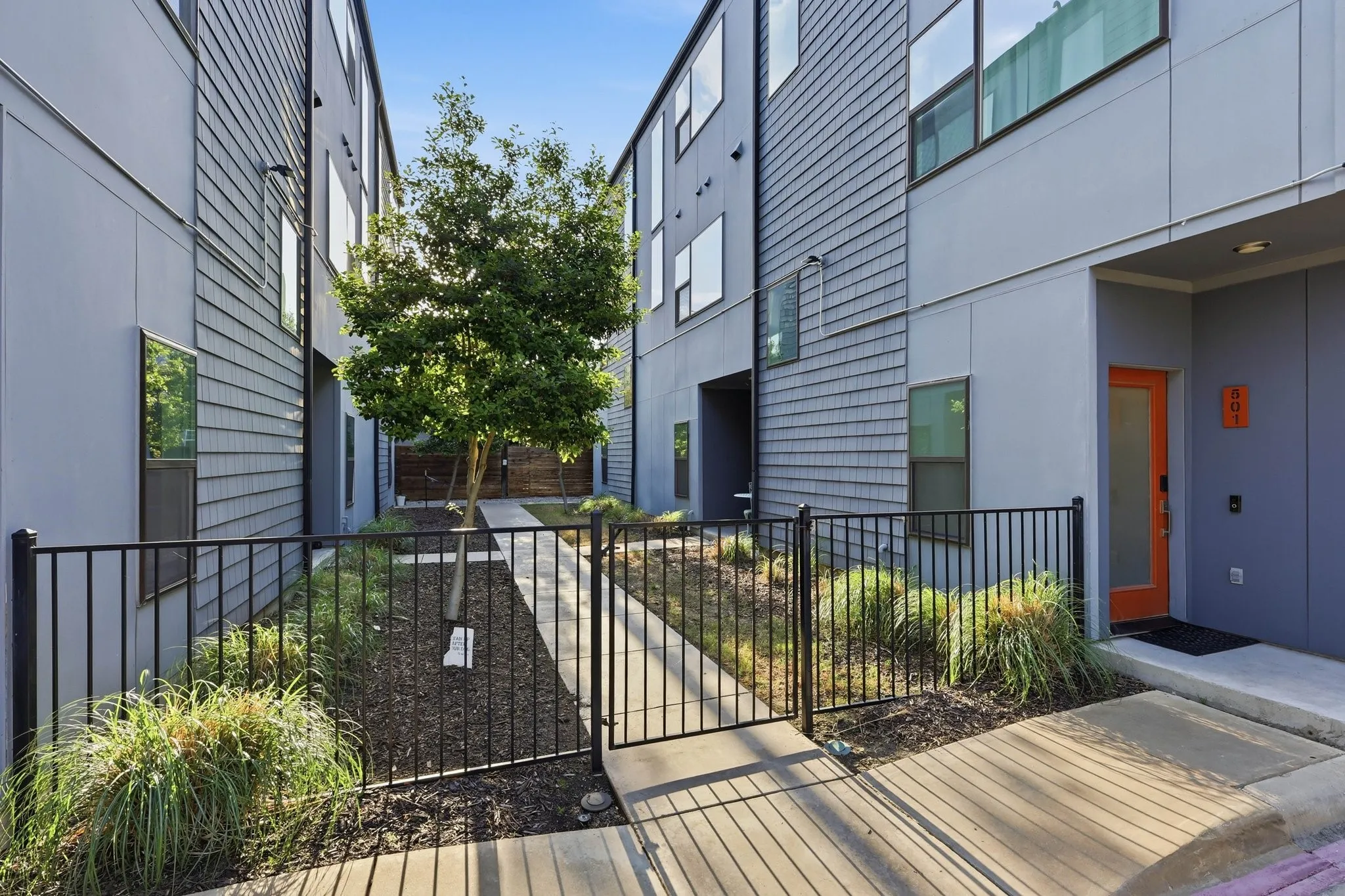 Modern townhome exterior with mixed material siding, featuring a vibrant orange entry door and black iron fencing enclosing a landscaped pathway