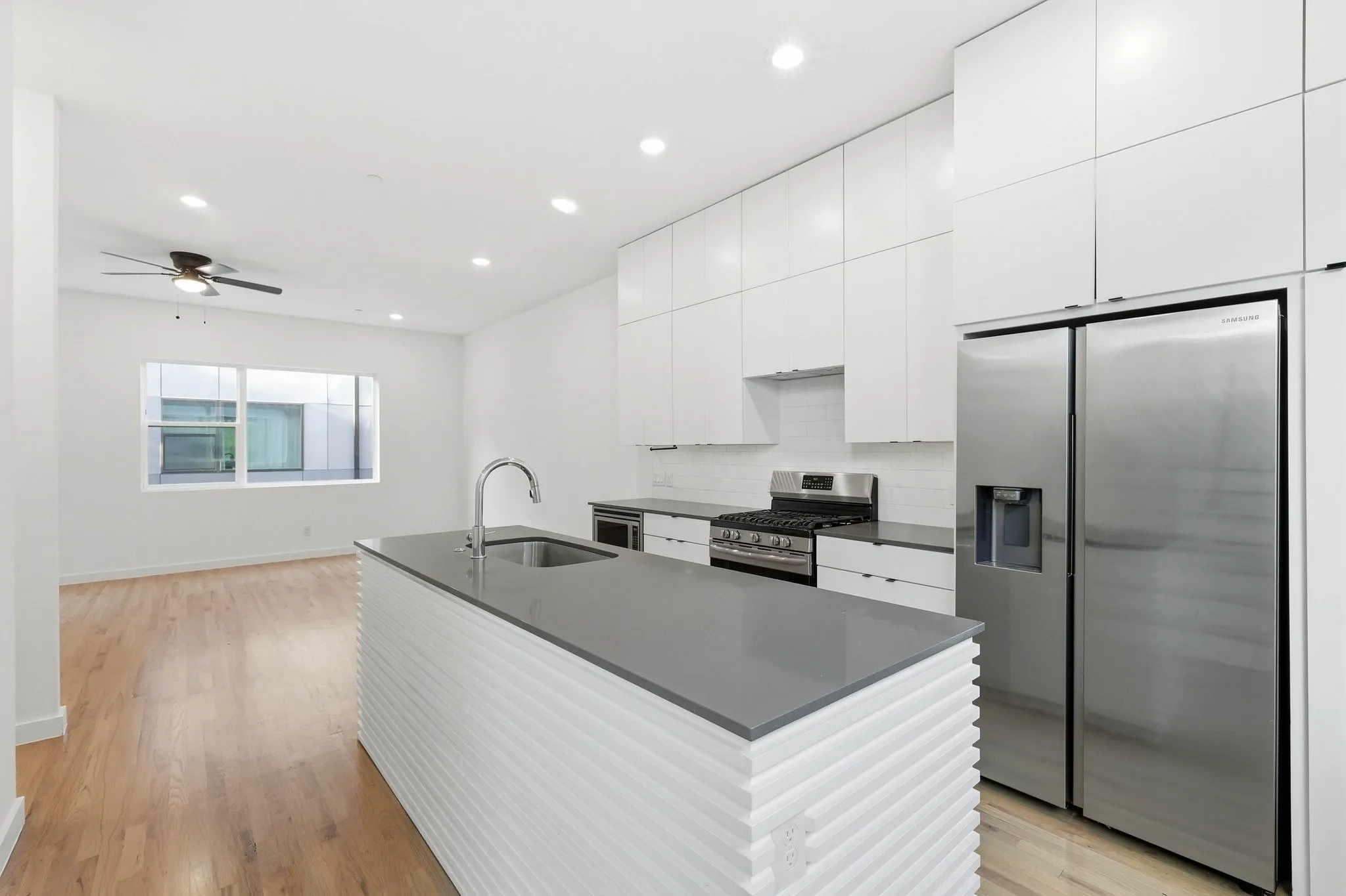 Modern kitchen featuring a central island with a grey countertop and integrated sink, stainless steel appliances, white flat-panel cabinetry, and wood-finish flooring