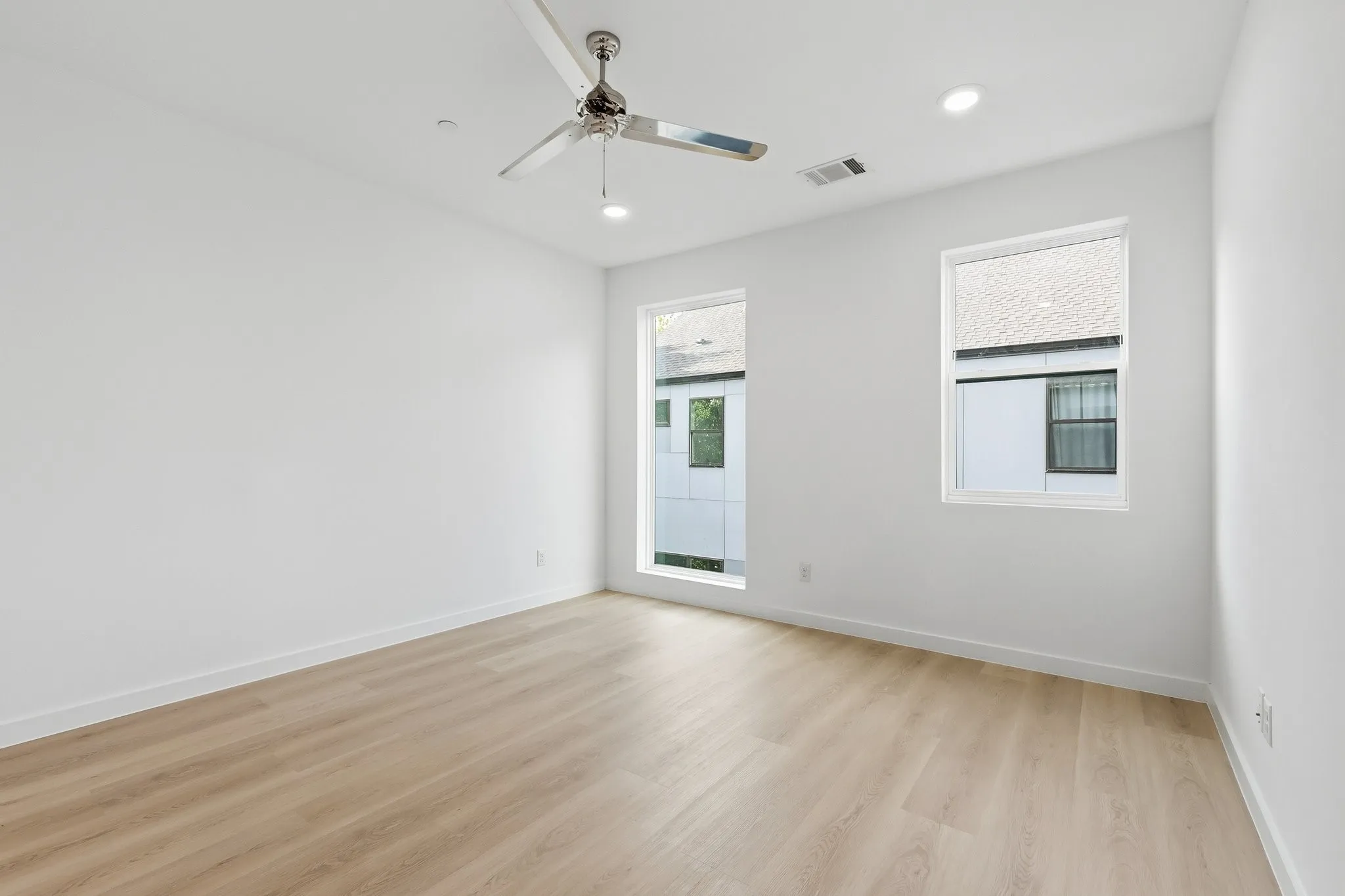 Light wood-finish flooring complements crisp white walls in this room featuring two windows with white trim, a ceiling fan, and recessed lighting
