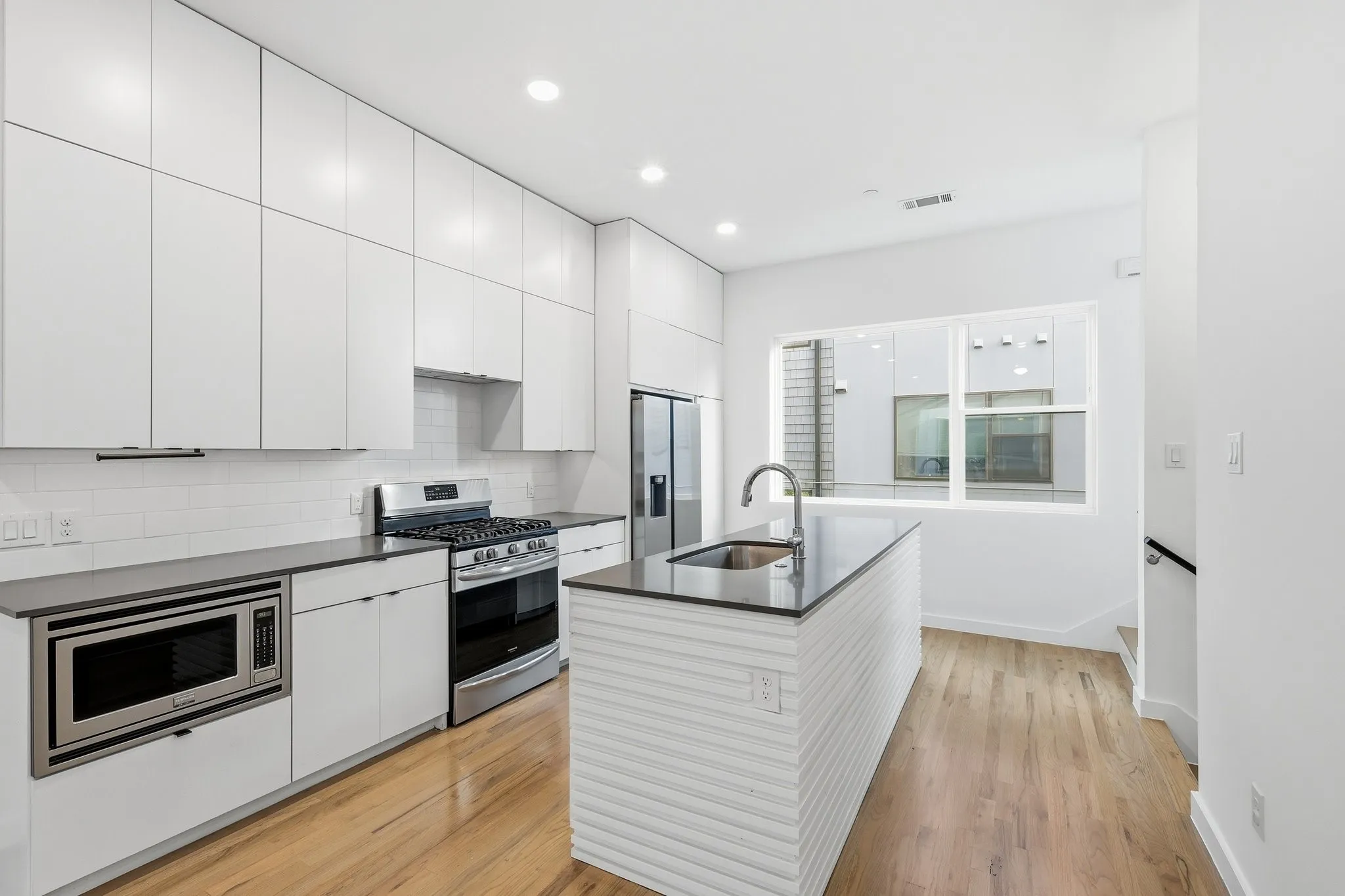 Modern kitchen featuring a central island with an undermount sink, stainless steel appliances, extensive white cabinetry, a subway tile backsplash, and light wood-finish flooring