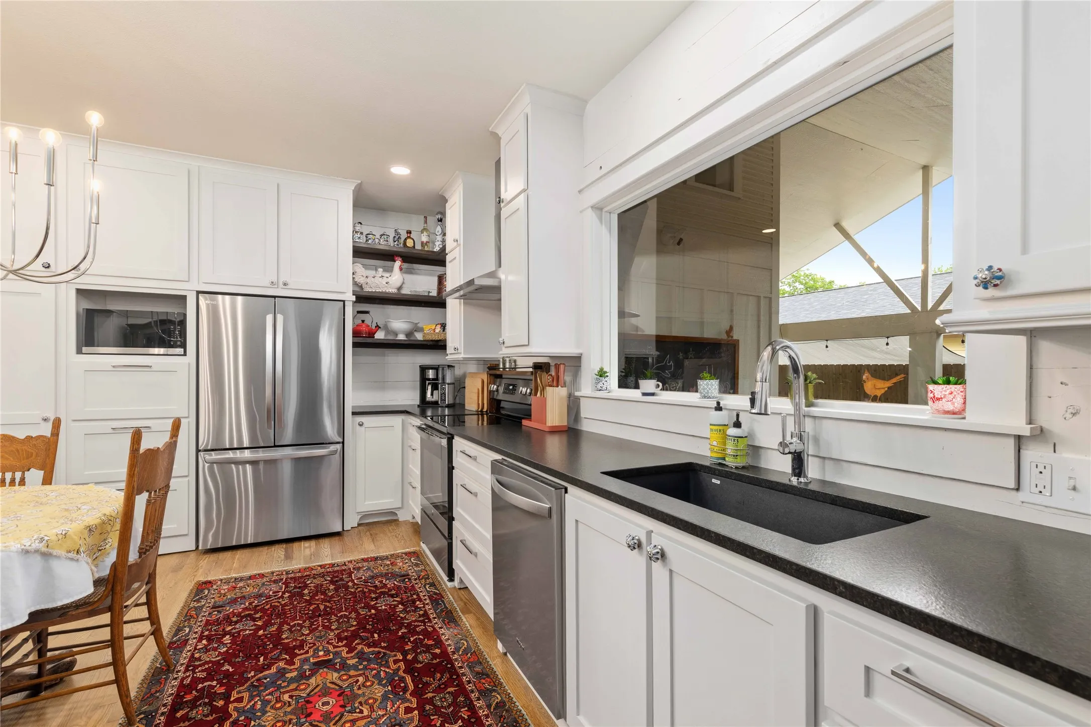 Modern kitchen featuring white shaker cabinetry, stainless steel appliances, dark countertops, and a large window overlooking an outdoor covered patio