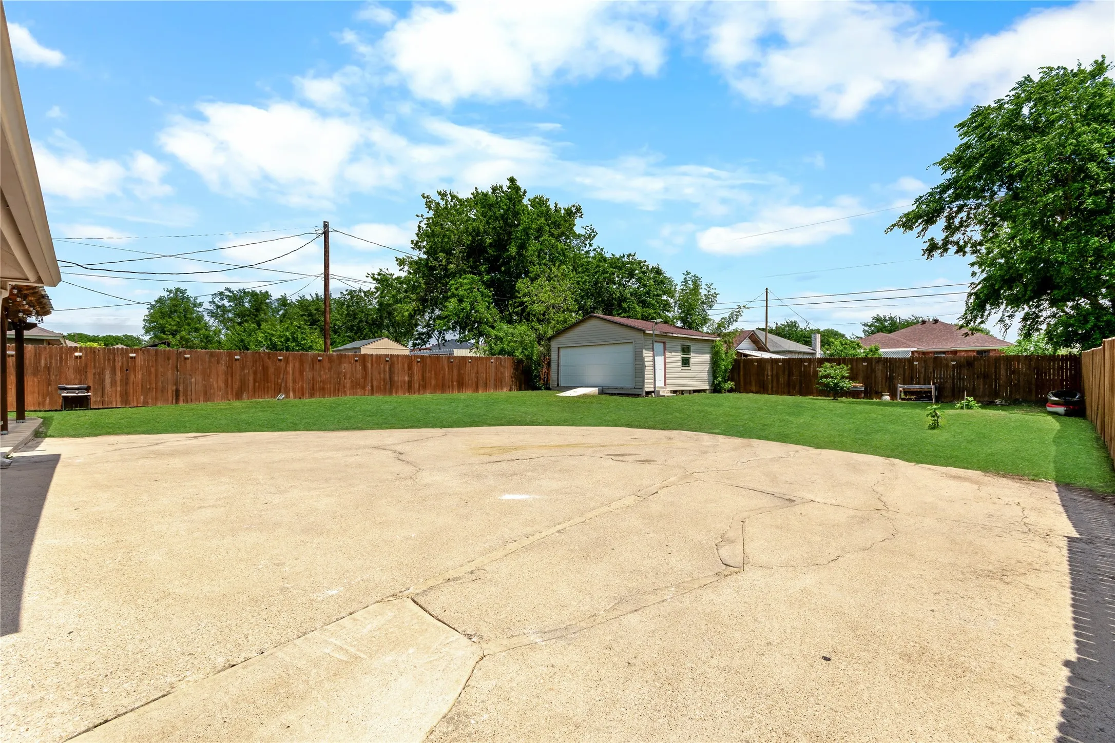 Expansive concrete patio area with a detached garage/workshop, lush green lawn, and privacy fencing