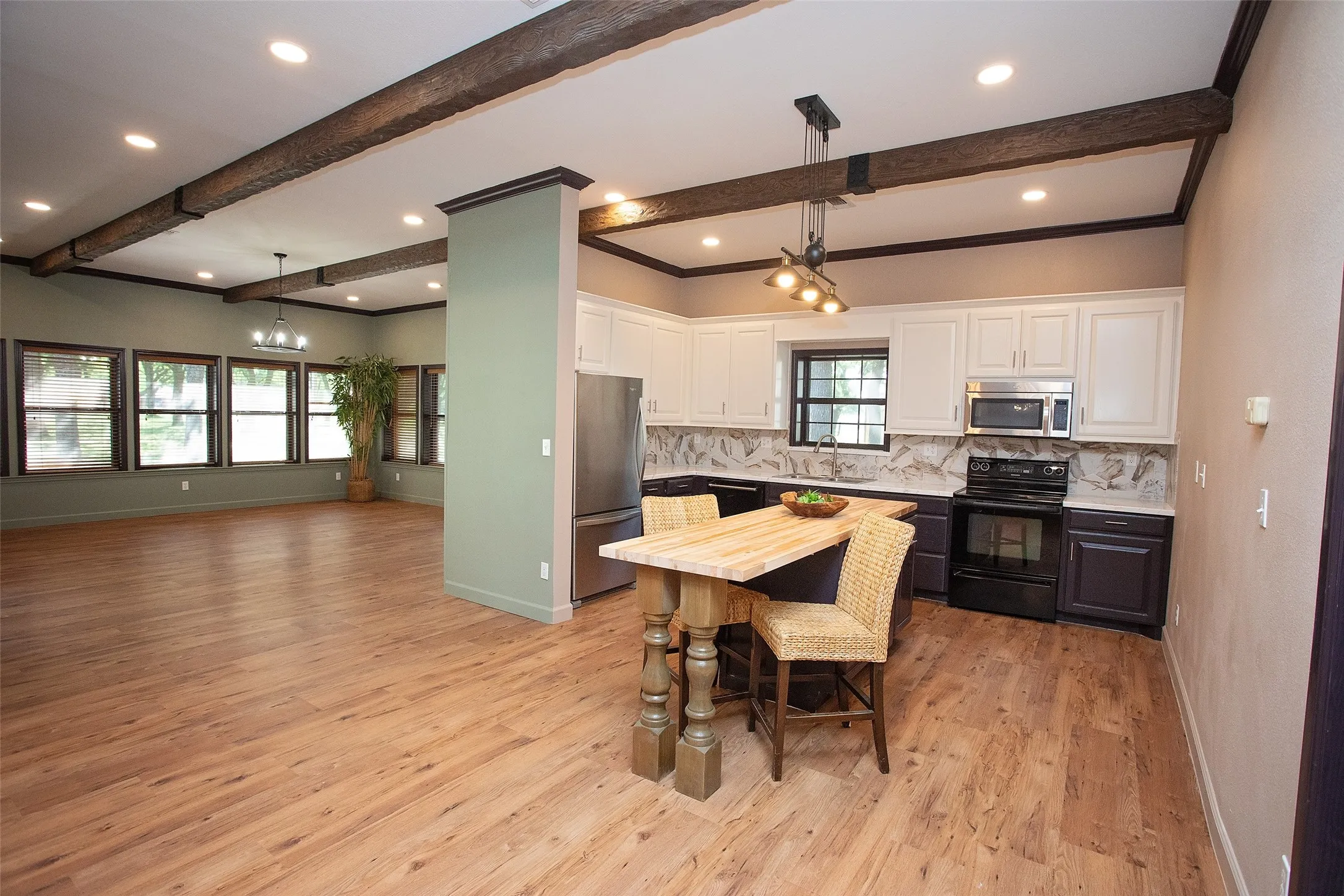 Open-concept kitchen, dining, and family room designed for seamless living and entertaining. The kitchen features ample cabinetry, stainless appliances, and a central island ideal for casual dining or gathering. Rich wood flooring and exposed beams carry throughout, adding warmth and character, while the expansive family room is lined with windows that bring in abundant natural light and showcase the scale of the space.