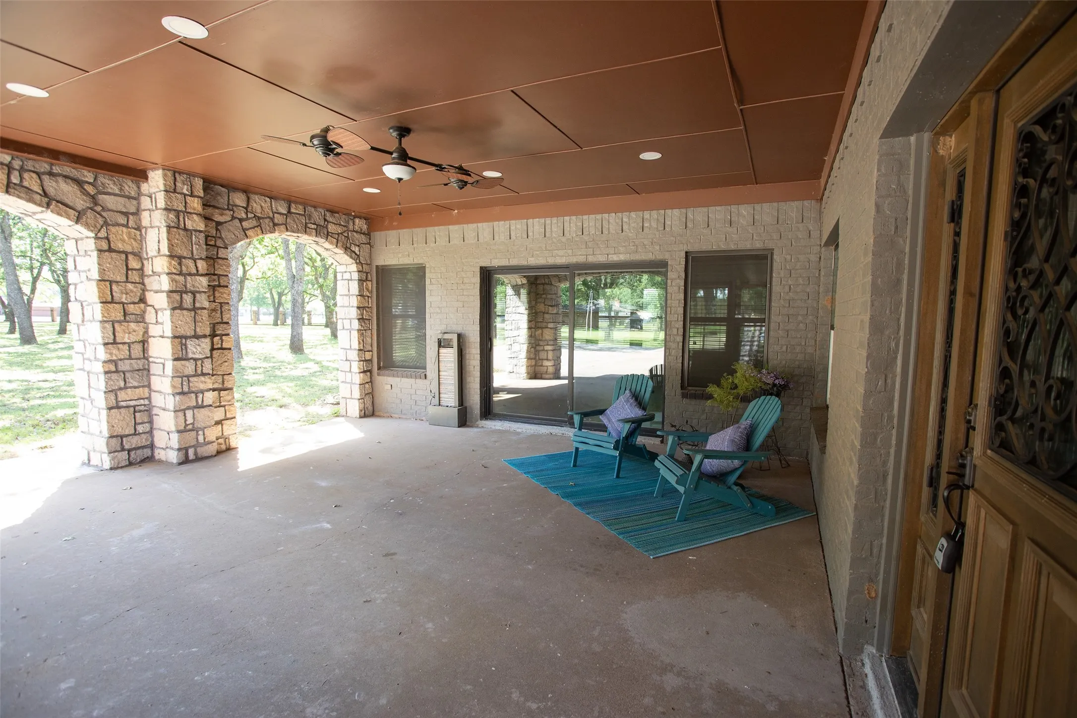 Covered front patio designed for relaxed outdoor living, featuring ample space for seating and entertaining. Framed by stone accents and accessed through sliding glass doors, this inviting area overlooks the front yard with mature trees, offering shade, privacy, and a peaceful setting to enjoy the outdoors.