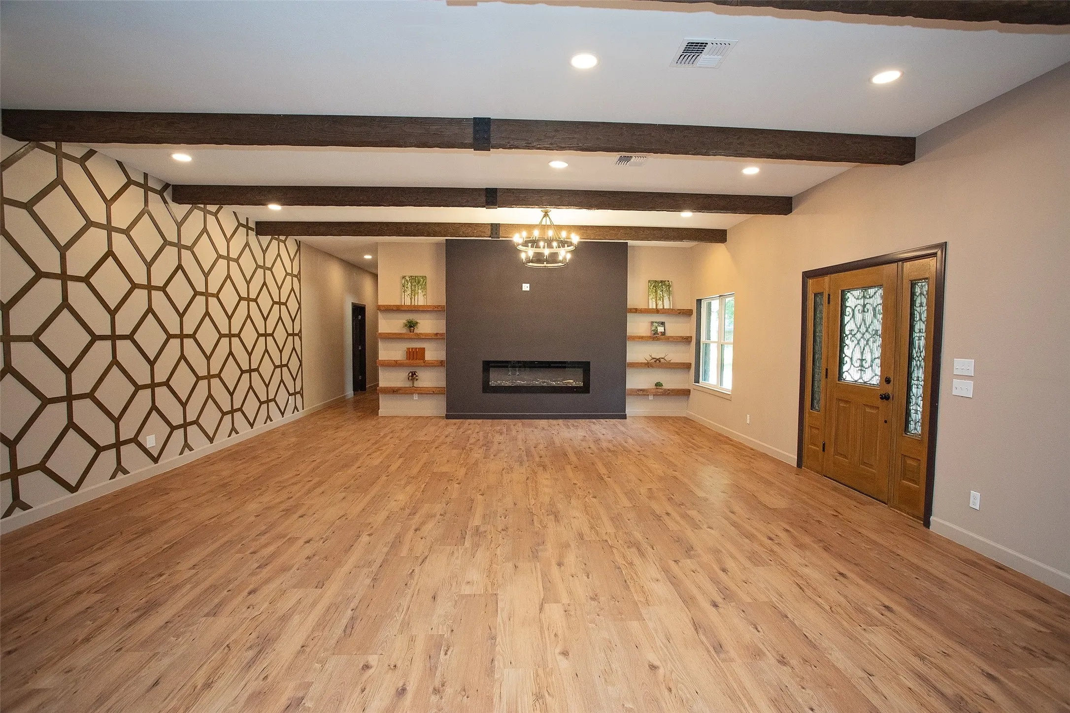 Spacious living room with an open layout designed for both everyday comfort and entertaining. Enter through the decorative front door into a welcoming space anchored by a sleek electric fireplace. Exposed wood beams add warmth and character, while the custom geometric accent wall creates a striking focal point. Built-in shelving offers both style and function, and the expansive floor plan provides flexibility for a variety of furniture arrangements.