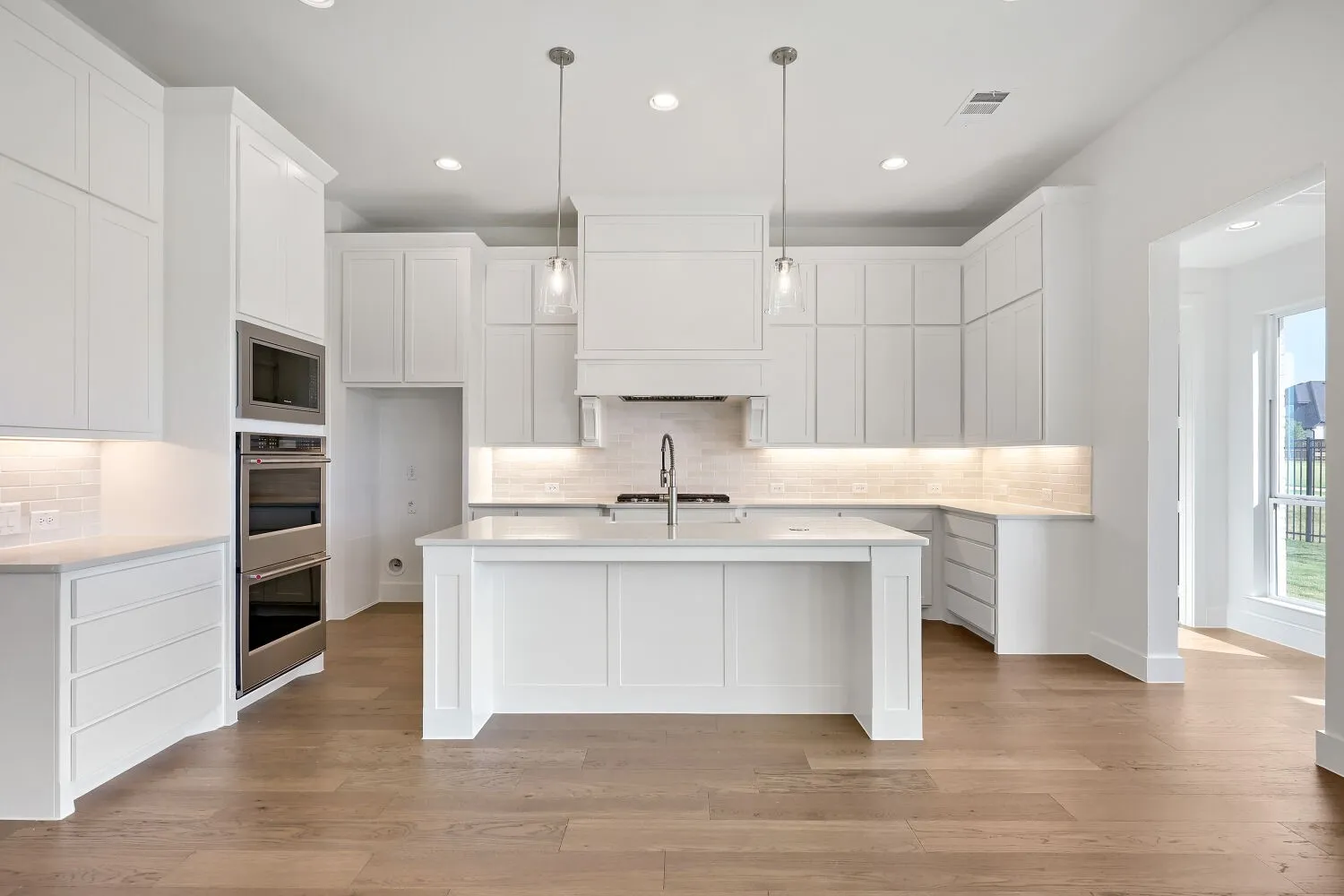 White cabinetry with under-cabinet lighting, stainless steel double ovens, central island with sink, subway tile backsplash, and wood-finish flooring