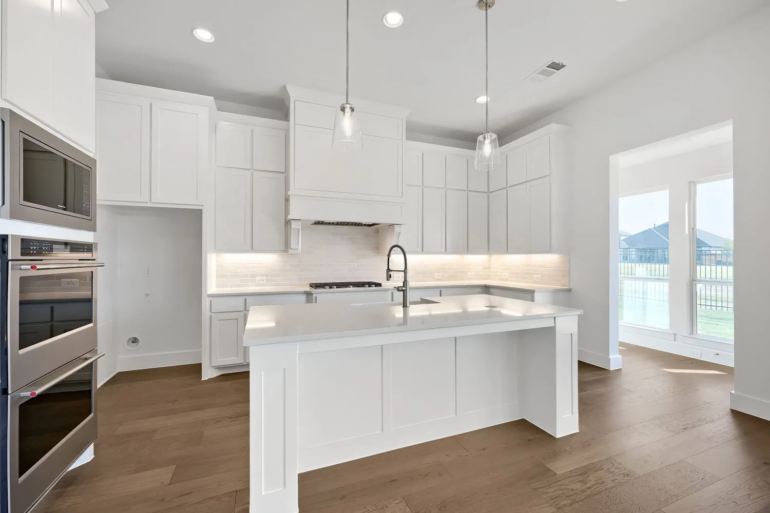 White cabinetry with undermount lighting, stainless steel double ovens, a central island with a sink, wood-finish flooring, and recessed ceiling lights