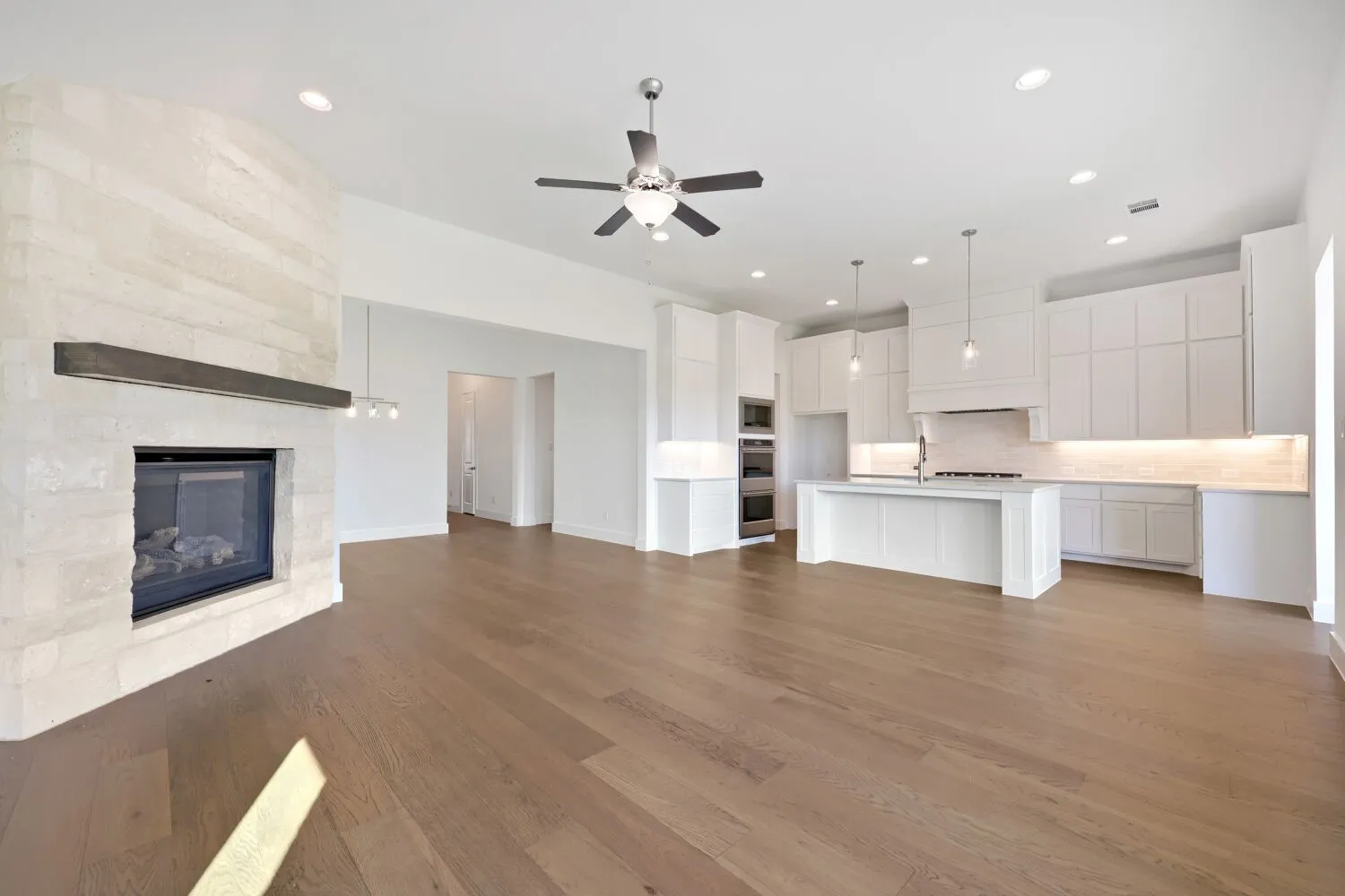 Stone-clad fireplace with mantel, wood-finish flooring, white kitchen cabinetry, kitchen island with sink, and recessed lighting