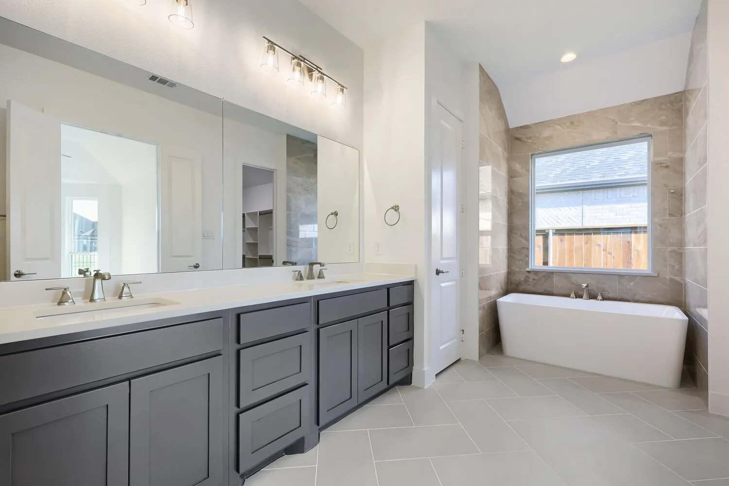 Dual vanity with undermount sinks and extensive countertop space, large wall-mounted mirror with overhead lighting, freestanding soaking tub, floor-to-ceiling tile surround, and light-toned tile flooring