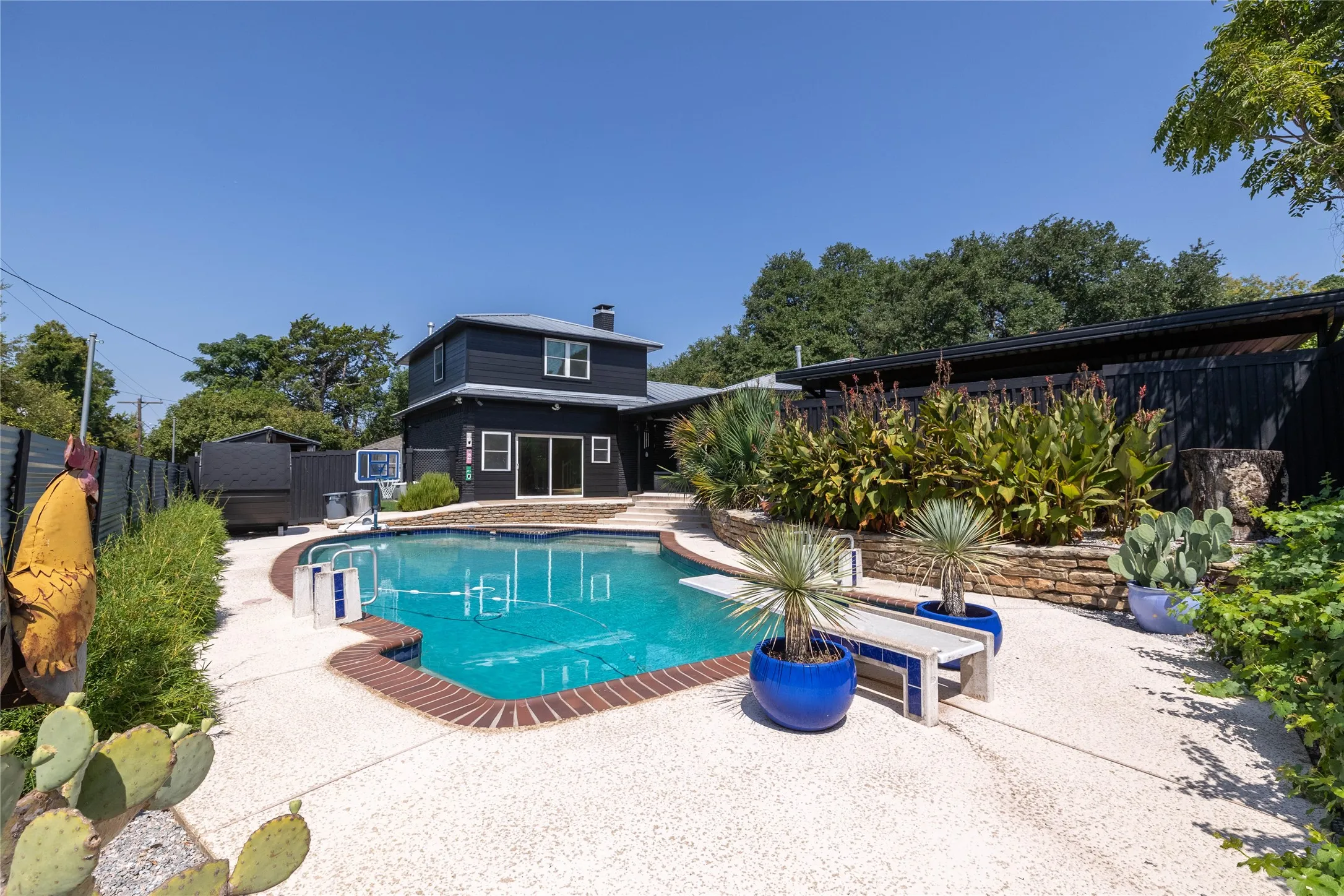 Resort-style backyard featuring a large swimming pool with brick coping, expansive white gravel decking, layered landscaping with mature plants, and a dark-painted exterior