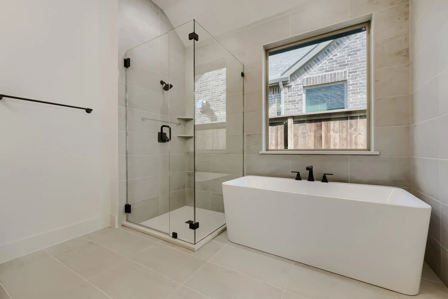 Freestanding soaking tub with matte black fixtures, glass-enclosed shower with tile surround and built-in shelving, and neutral tile flooring