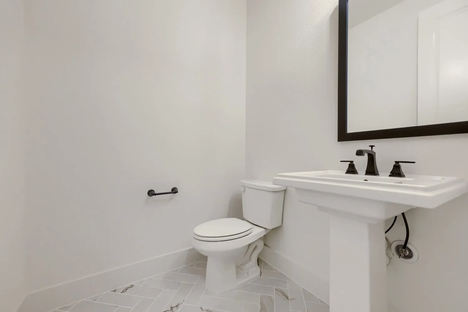 Pedestal sink with dark bronze fixtures, framed mirror, and light-toned tile flooring in a herringbone pattern