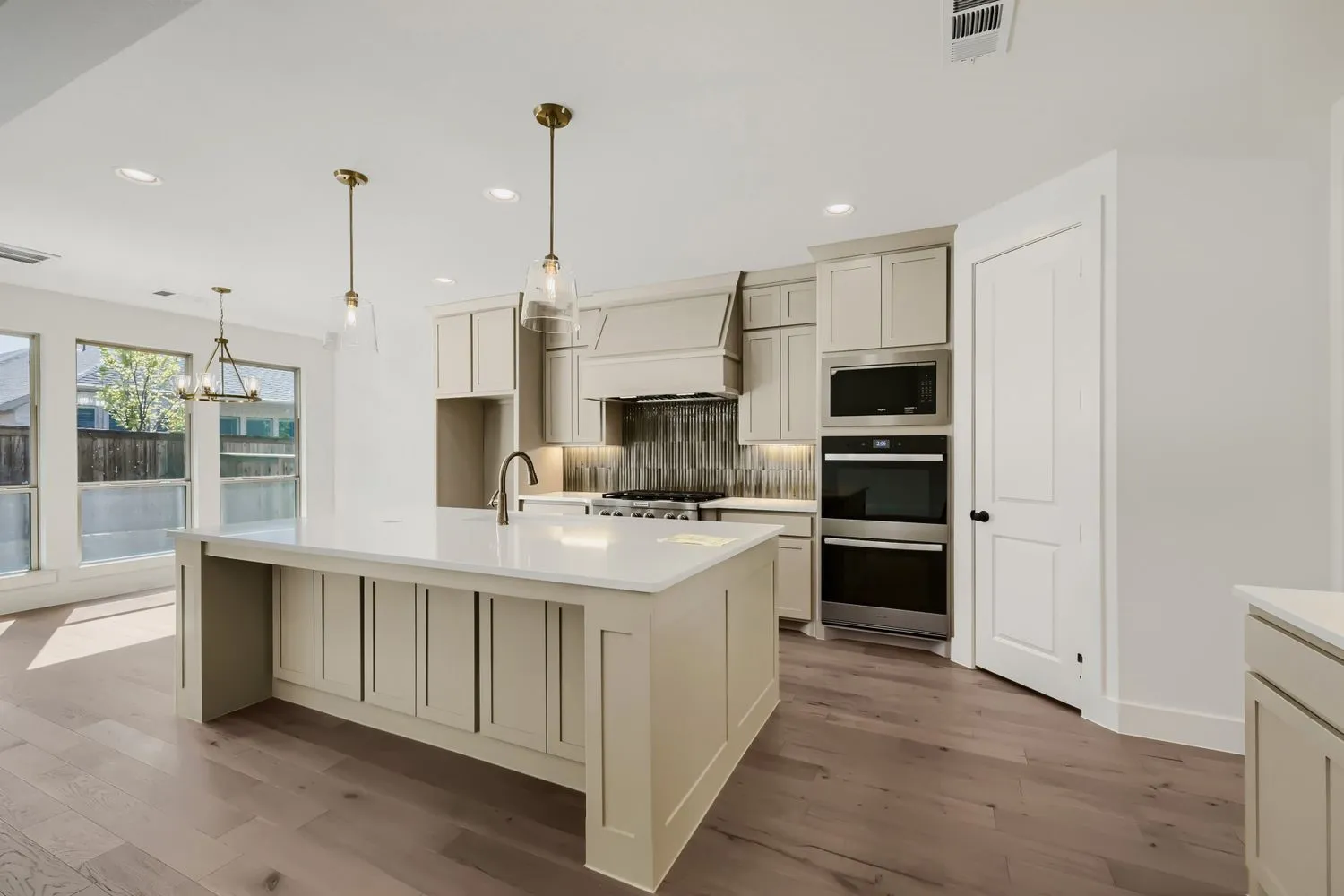 Kitchen featuring an island with a built-in sink, stainless steel appliances, a tile backsplash, light-toned cabinetry, and wood-finish flooring