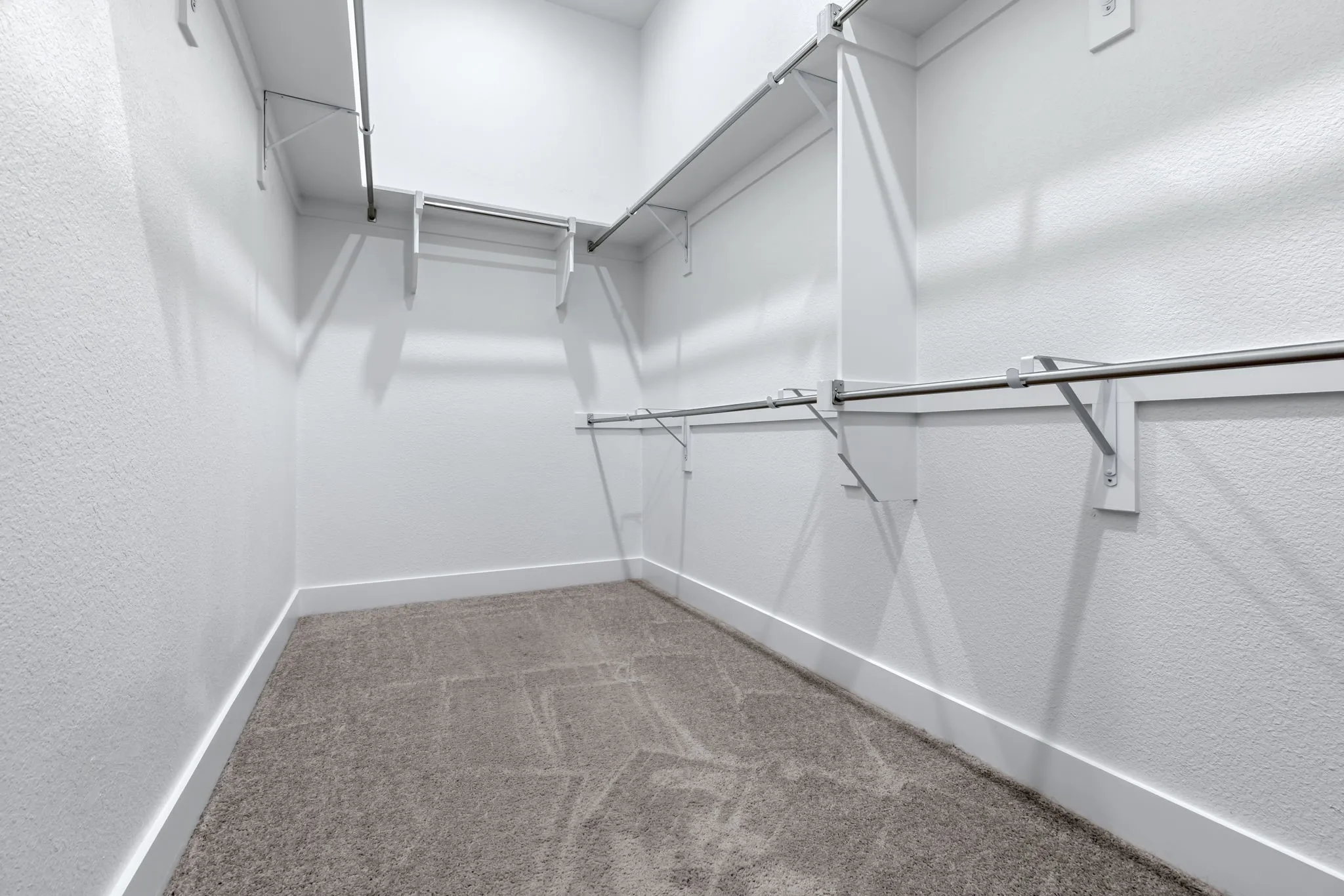 Spacious walk-in closet featuring light gray patterned carpet, white baseboards, and built-in white shelving with chrome hanging rods