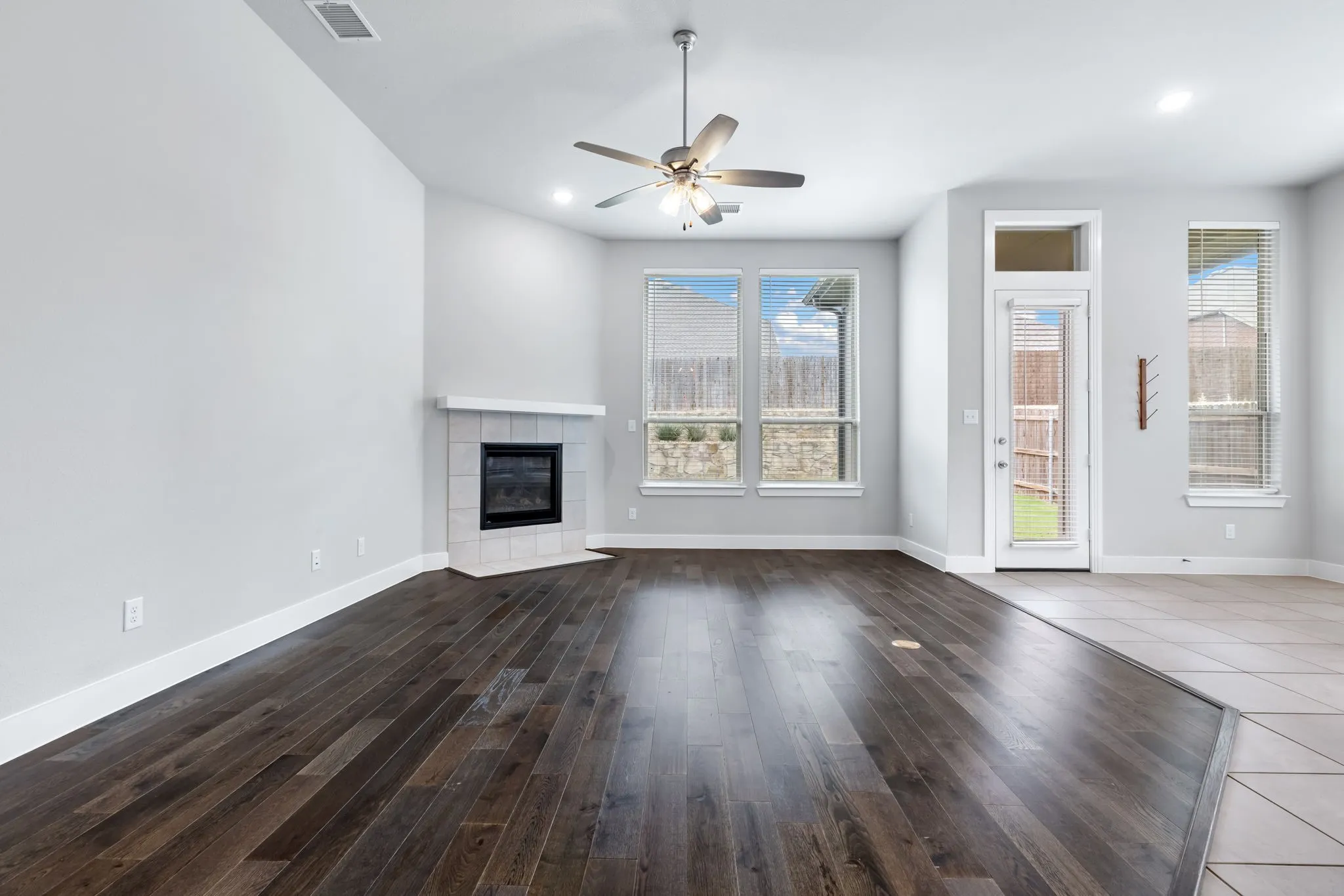 Spacious living area featuring dark wood-finish flooring, a corner fireplace with a white tile surround, and multiple windows
