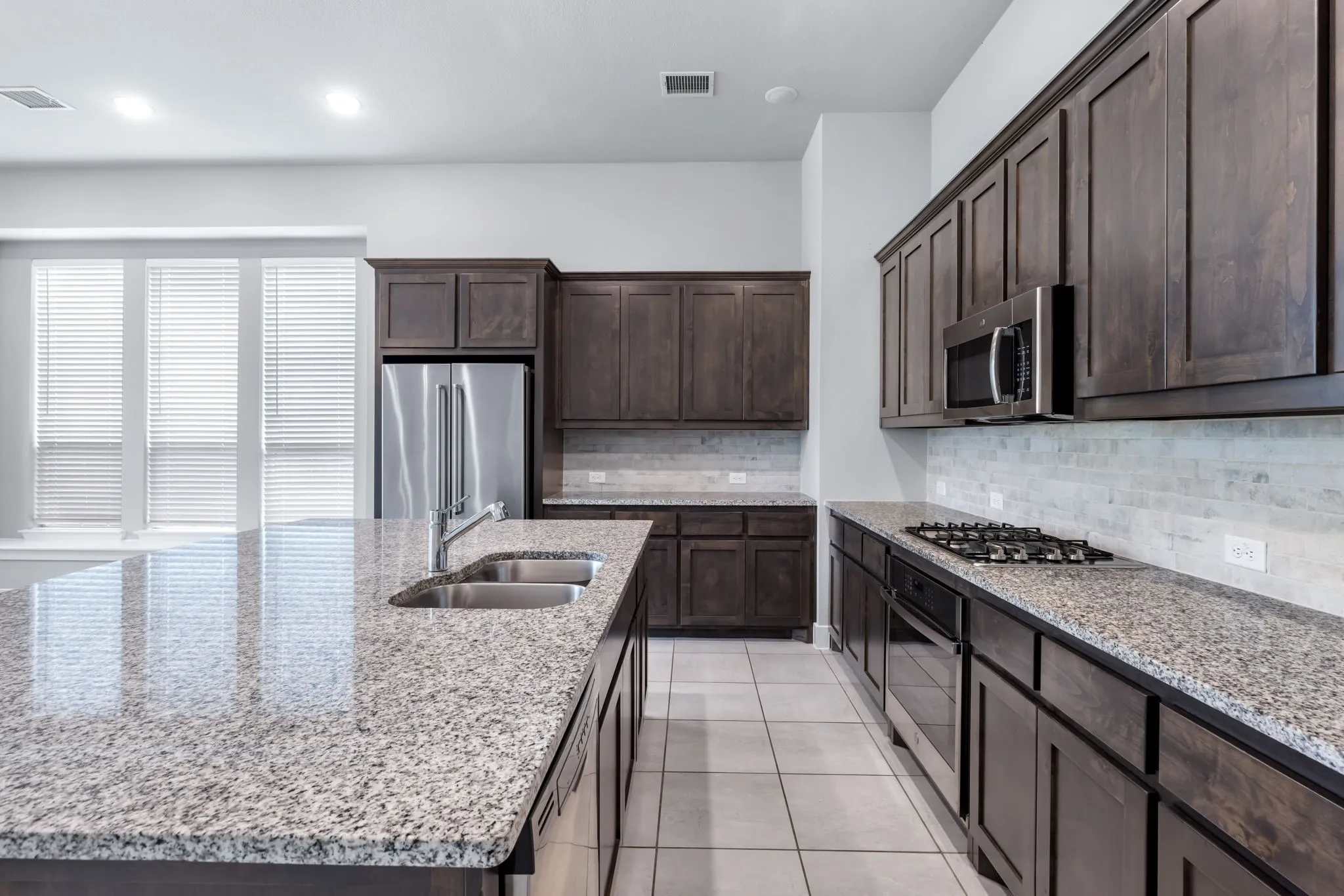 Kitchen featuring an expansive island with a double basin sink, stainless steel appliances, dark wood-finish cabinetry, speckled countertops, and a subway tile backsplash