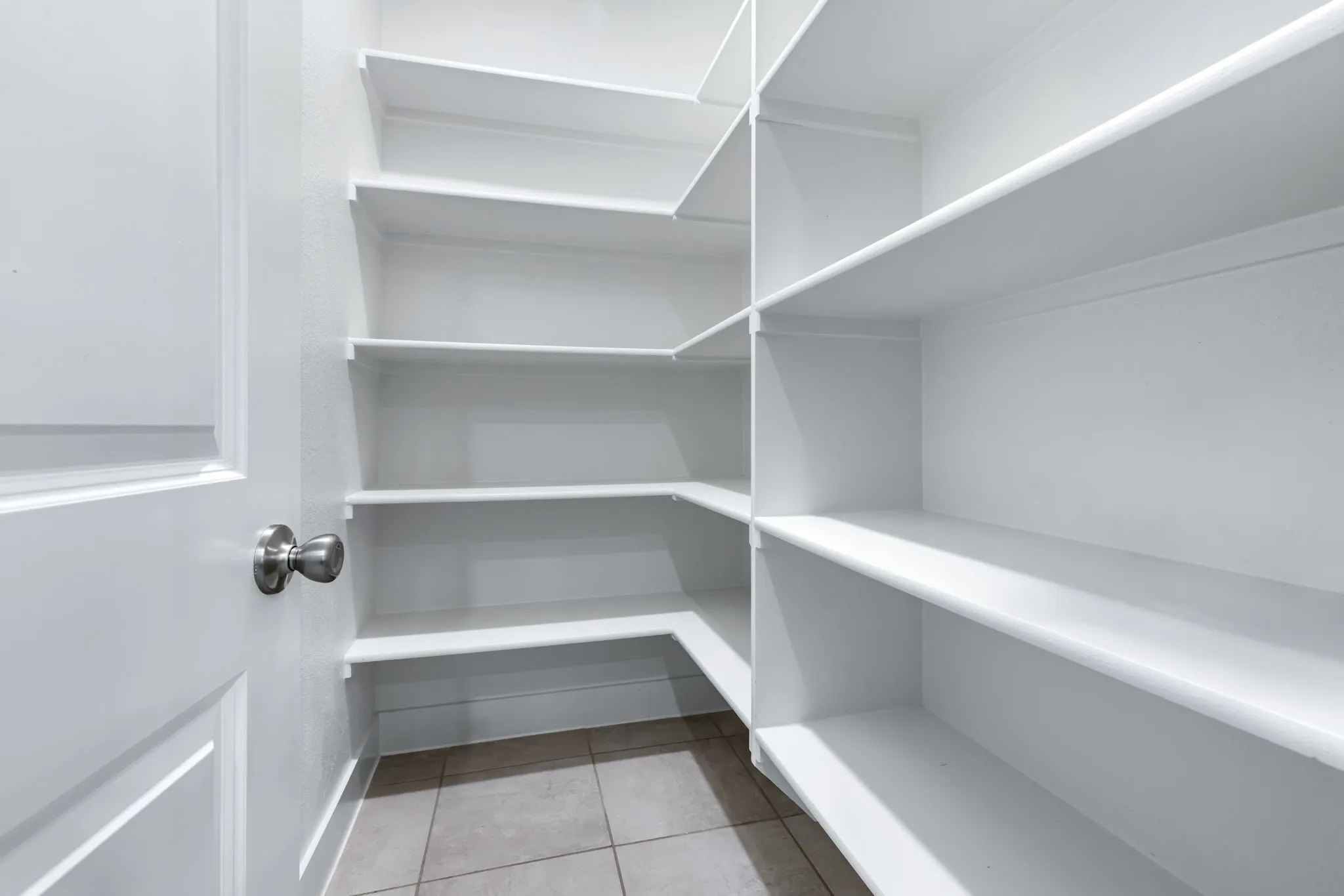 Walk-in pantry featuring extensive white shelving, tiled flooring, and a paneled door with a spherical knob