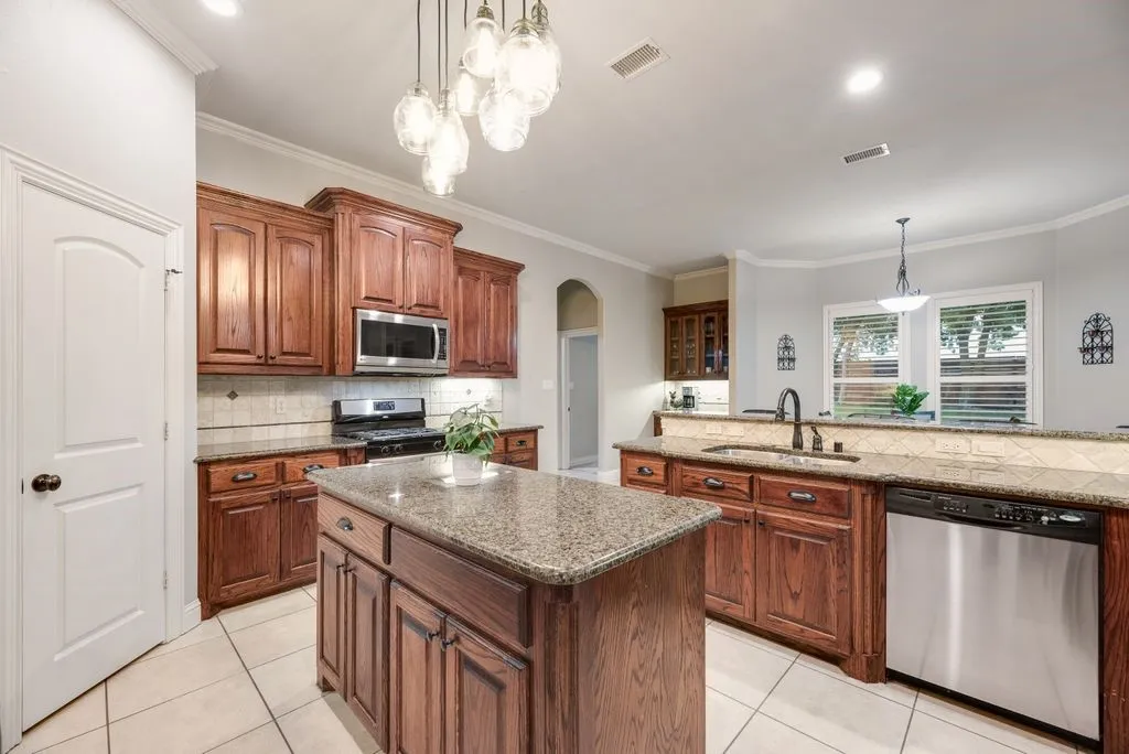 Kitchen featuring a central island with a granite-style countertop, tile flooring, rich wood cabinetry, stainless steel appliances, and a decorative pendant light fixture