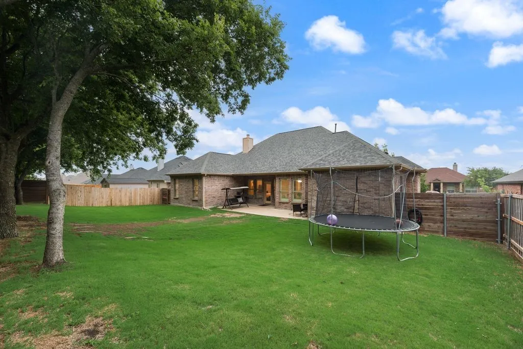 Expansive backyard featuring a manicured lawn, established shade trees, a red brick exterior, a covered patio, and a privacy fence