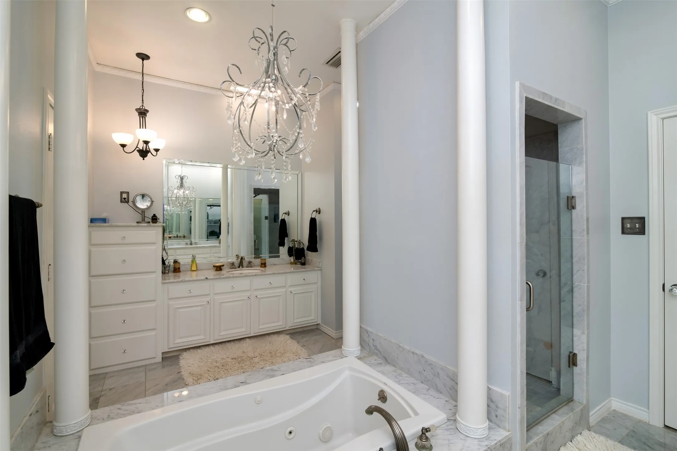 Contemporary bathroom featuring a jetted tub set in a marble surround, a dual vanity with white cabinetry and light-toned countertops, a glass-enclosed shower with marble-look tile, and decorative chandeliers