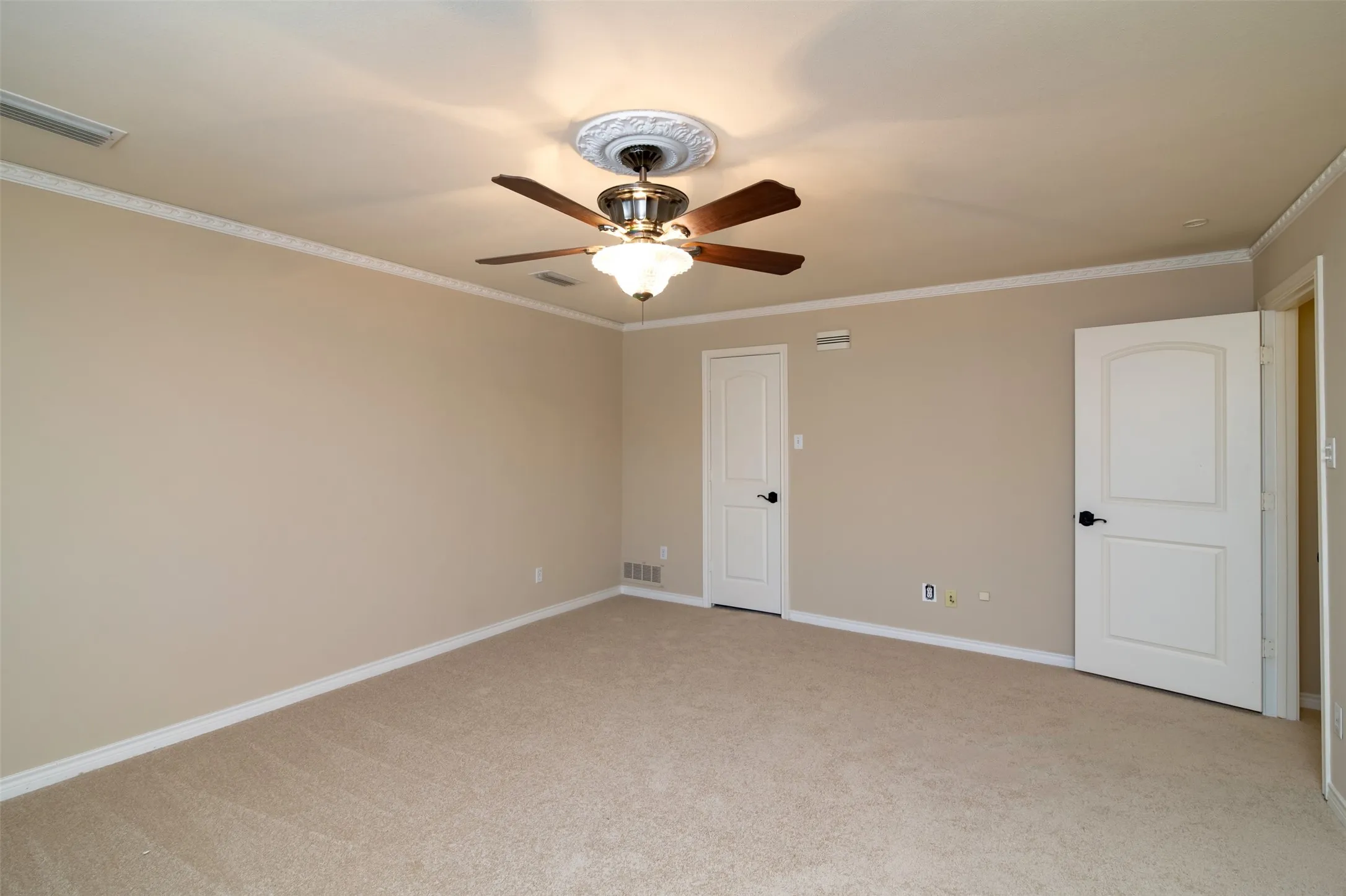 Spacious room featuring neutral carpet flooring, light beige wall paint, white crown molding, and a decorative ceiling fan with a medallion