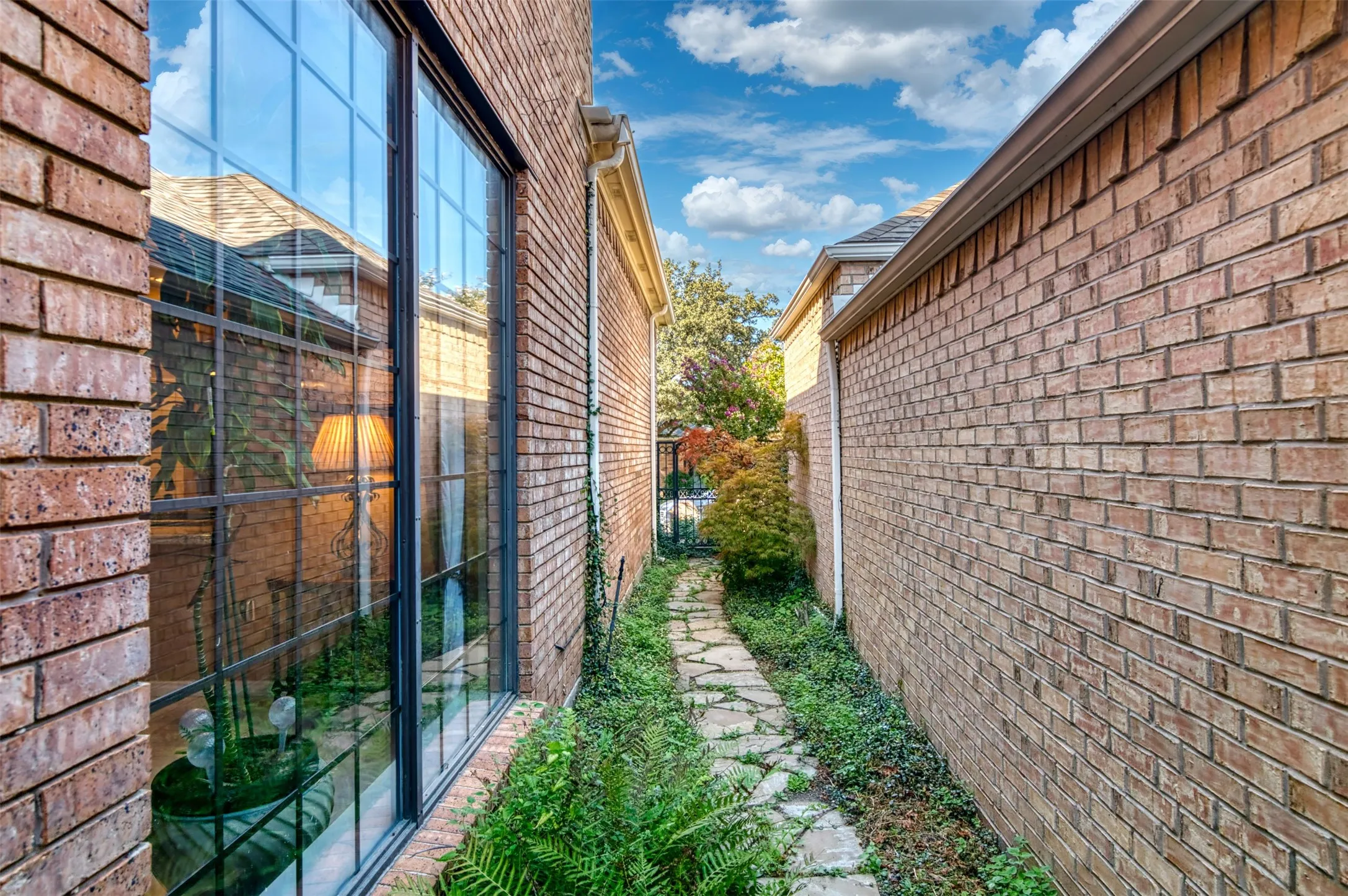 Stone pathway bordered by mature plantings, featuring brick exterior walls and a large, multi-pane window