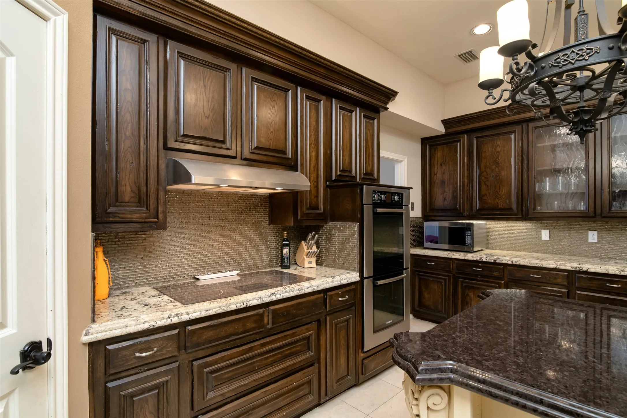 Kitchen featuring dark wood cabinetry, granite countertops, a built-in oven, a stainless steel range hood, and a mosaic tile backsplash