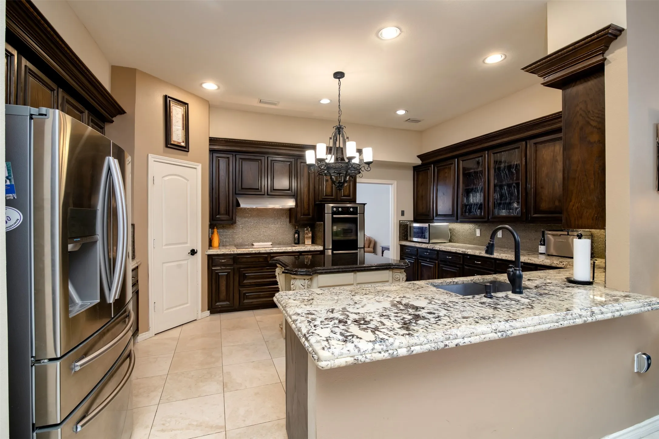 Gourmet kitchen featuring dark wood cabinetry, light granite countertops, a central island with an additional sink, stainless steel appliances, and recessed lighting