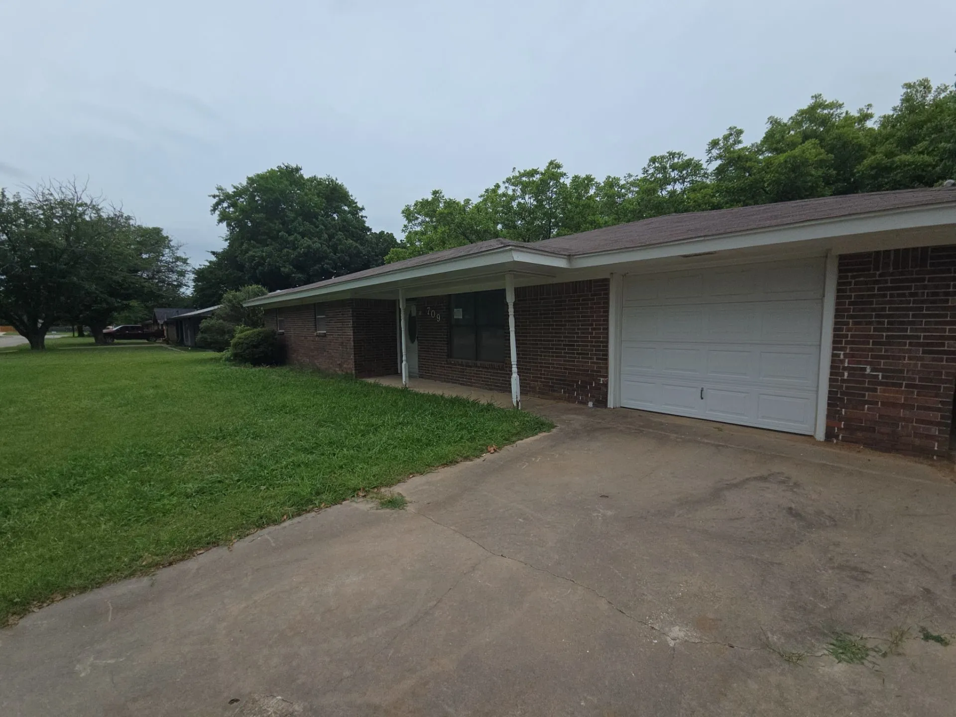 Brick exterior home featuring a one-car garage, covered entry, and a spacious front lawn with mature trees